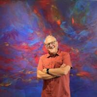 Halbkörperportrait von Stefan Ruppaner, der mit freundlichem Lächeln und verschränkten Armen vor einer abstrakt bunt bemalten Wand steht.