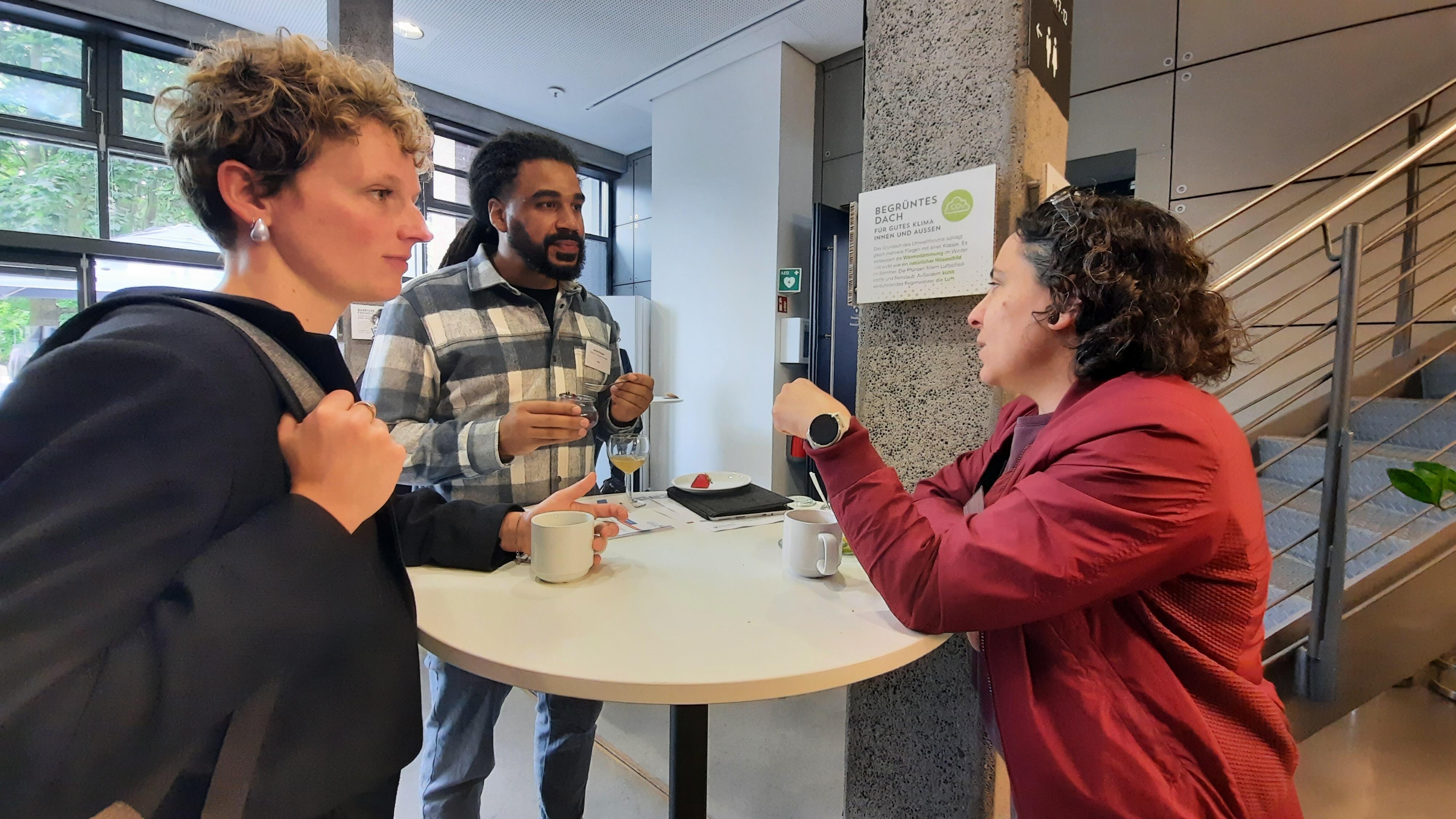 Drei Teilnehmende, zwei Frauen und ein Mann, stehen an einem runden Stehtisch im Gespräch.