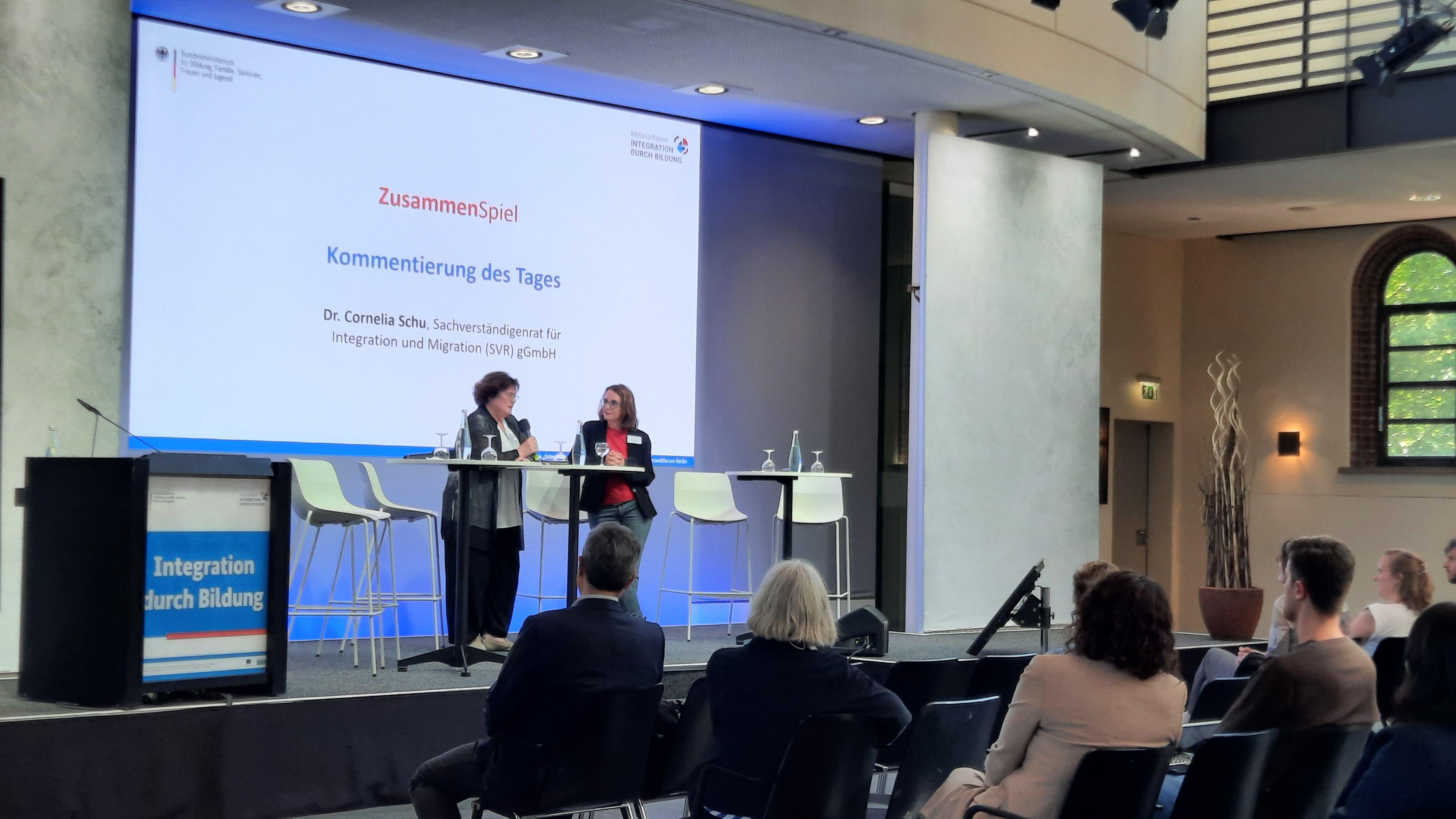 Prof. Dr. Dr. h.c. Ingrid Gogolin und Dr. Cornelia Schu stehen an einem Stehtisch auf dem Podium der Vernetzungstagung im Gespräch.