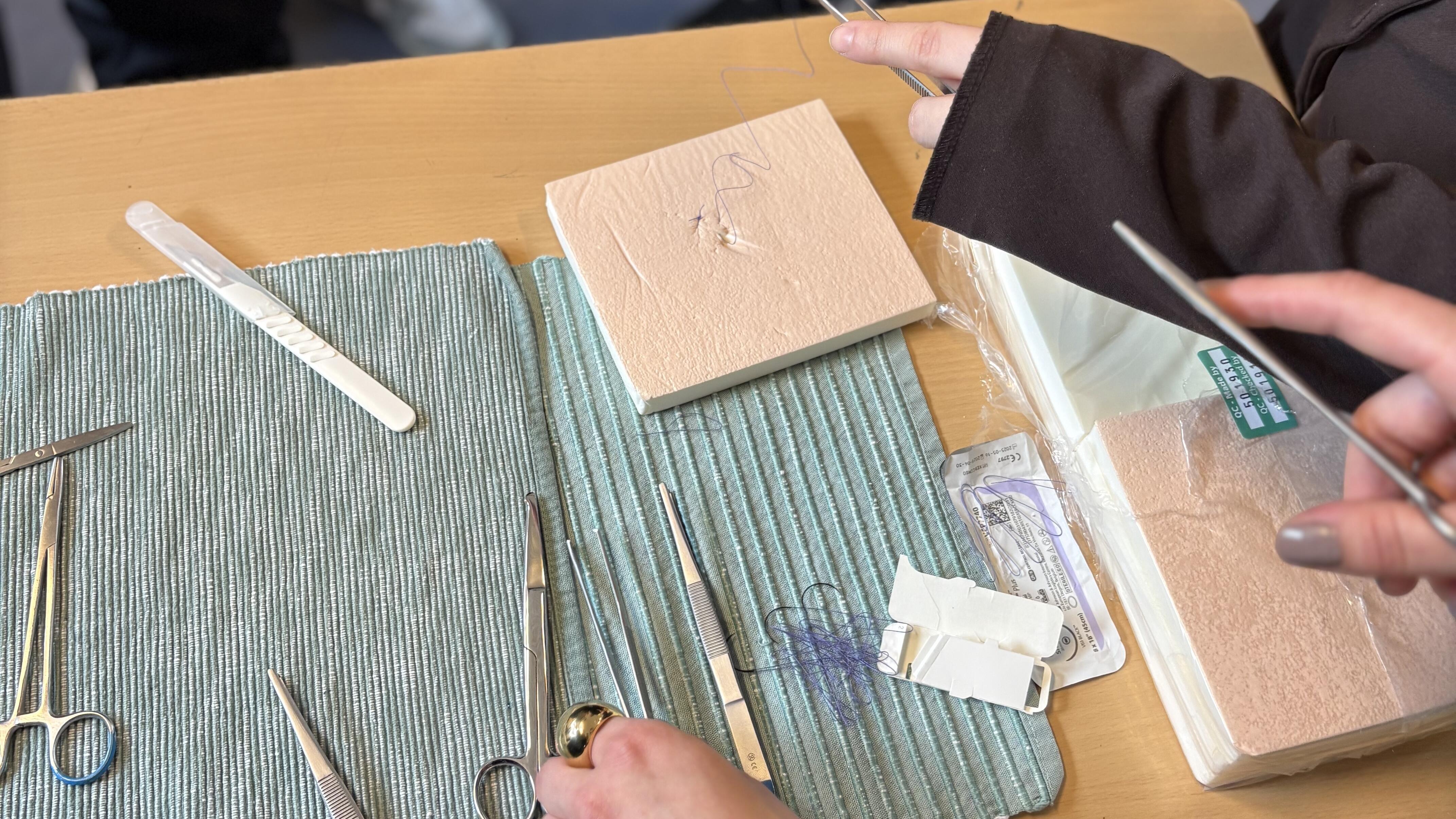 Auf einem Tisch liegen chirurgische Nadeln, Nahtfäden und eine Übungsplatte mit Beispielnähten zum Üben von Wundverschlüssen.