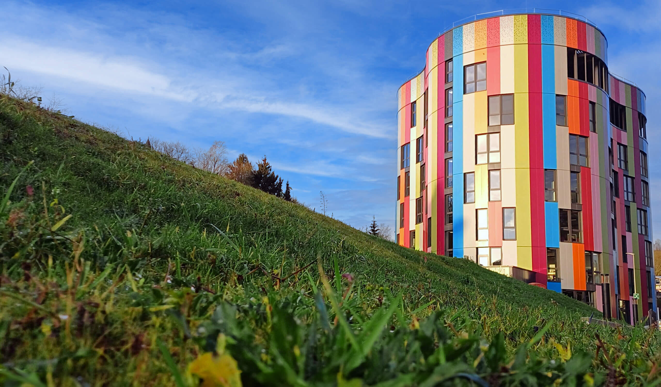 Außenansicht des bunten Hauptgebäudes bei Tag mit Wiesenhügel im linken Bildvordergrund.