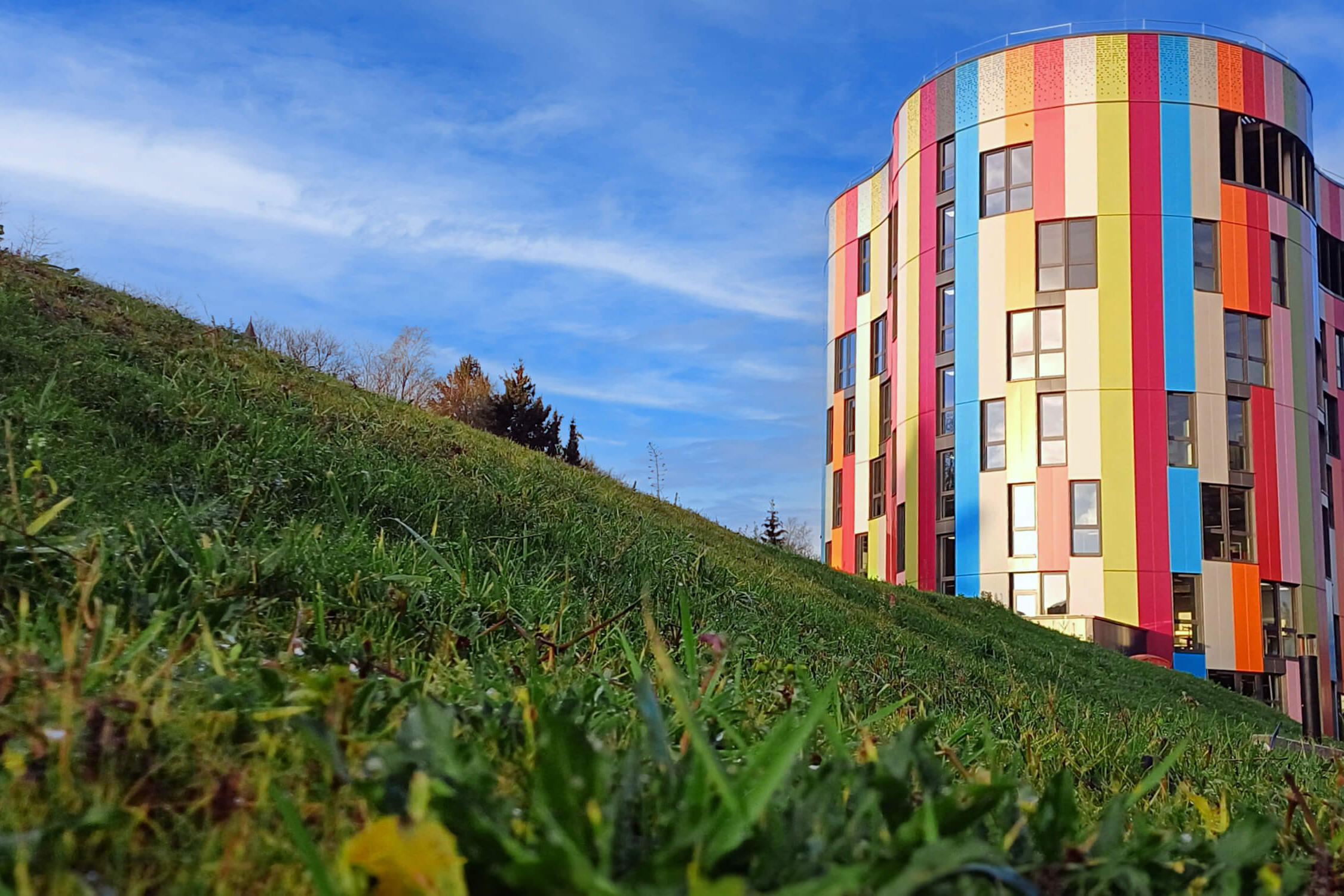 Außenansicht des bunten Hauptgebäudes bei Tag mit Wiesenhügel im linken Bildvordergrund.