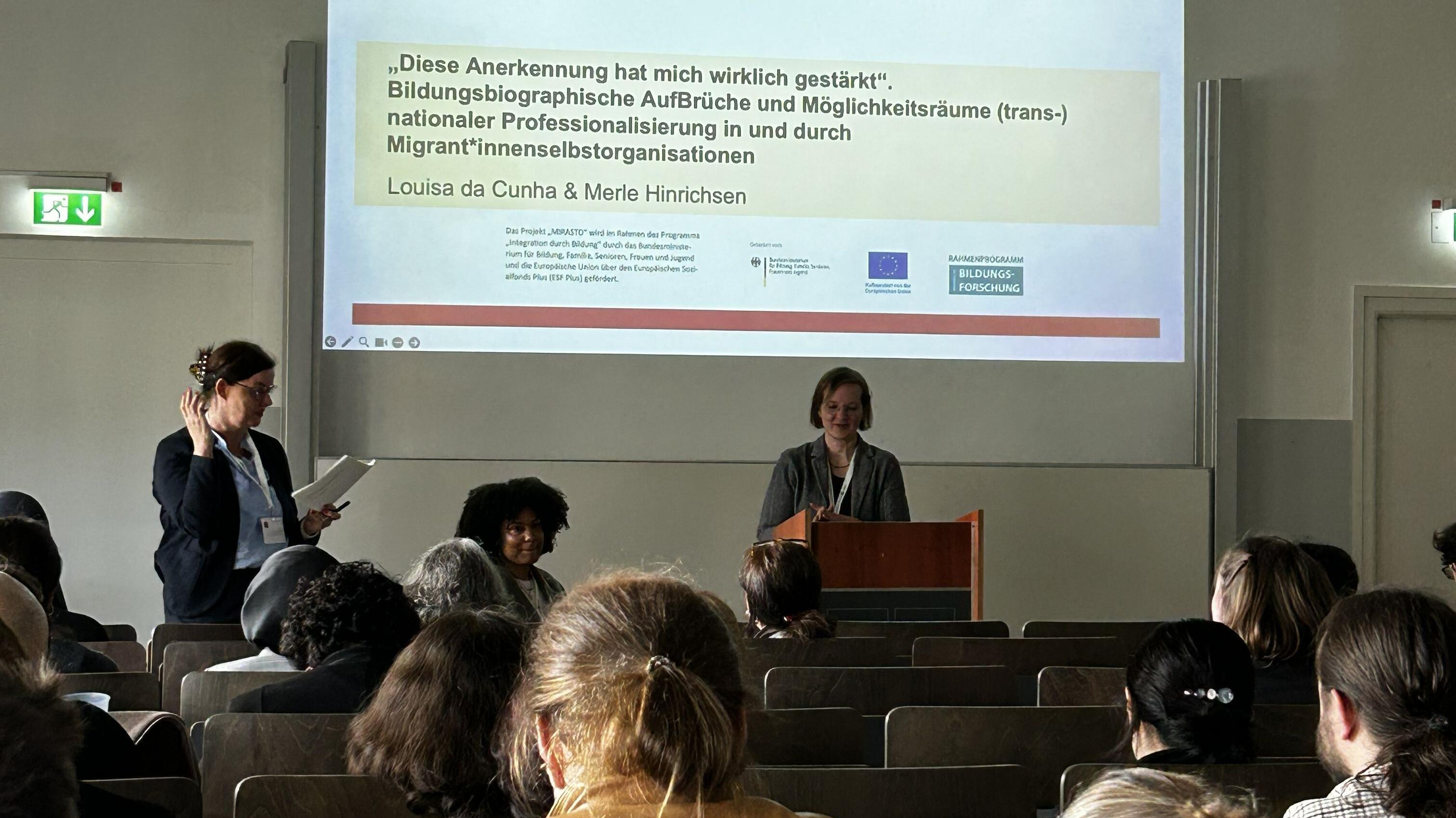 Louisa da Cunha und Merle Hinrichsen halten eine Präsentation im Saal.