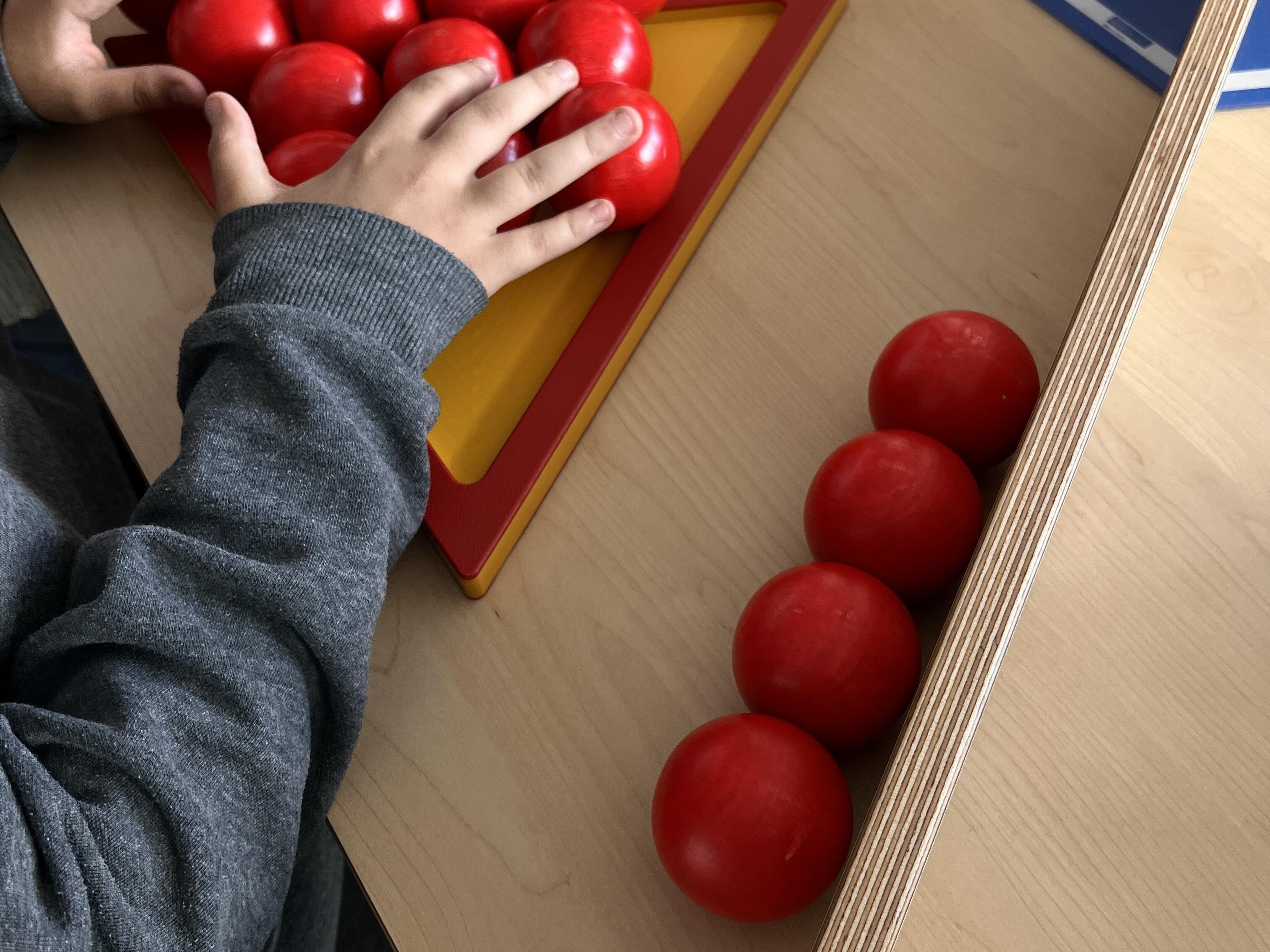 Kurzbeschreibung:
Hände ordnen rote Kugeln auf einem dreieckigen Spielfeld auf einem hellen Holztisch an. Daneben liegen weitere rote Kugeln und eine blaue Informationskarte.

Ausführliche Bildbeschreibung:
Auf einem hellen Holztisch befindet sich ein dreieckiges Spielfeld mit rotem Rand und gelber Innenfläche. Darauf liegen viele glänzende rote Kugeln dicht beieinander. Kleine Hände schieben oder sortieren die Kugeln auf dem Spielfeld.

Rechts neben dem Dreieck liegen vier weitere rote Kugeln in einer Reihe auf dem Tisch. Oben im Bild ist eine weitere Hand zu sehen, die über das Spielfeld greift. Im Hintergrund liegt eine blaue Informationskarte mit Text, der nur teilweise lesbar ist.

Das Bild zeigt eine interaktive Mitmachstation, bei der Kugeln auf einer geometrischen Fläche angeordnet werden. Der Fokus liegt auf dem Ausprobieren mit den Händen und den auffälligen roten Kugeln.

(Bildbeschreibung mit KI generiert.)