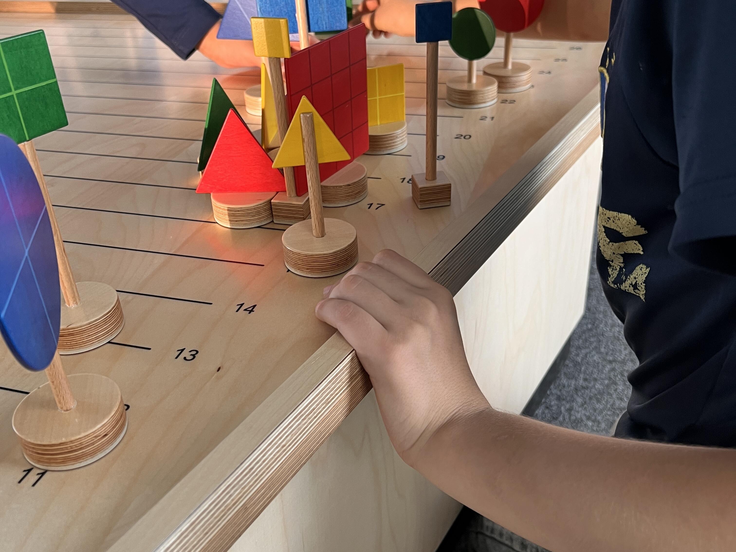 Kurzbeschreibung:
Ein Kind steht an einer Mitmachstation und ordnet bunte geometrische Holzformen auf einem Spielfeld an. Die Formen erinnern an Häuser, Bäume und Schilder.

Ausführliche Bildbeschreibung:
Auf einem hellen Holztisch stehen viele bunte Holzformen auf kleinen runden Sockeln. Zu sehen sind Dreiecke, Quadrate, Kreise und Rechtecke in Rot, Blau, Grün und Gelb. Die Formen sind auf einem Spielfeld mit Linien und Zahlen angeordnet und ergeben zusammen eine kleine Stadt- oder Landschaftsszene.

Rechts steht ein Kind am Tisch und hält sich am Rand fest. Eine weitere Person greift von links in die Szene und bewegt eine blaue Form. Im Hintergrund ist eine Wandgestaltung mit stilisierten Häusern, Bäumen und Türmen zu sehen.

Das Bild zeigt eine interaktive Lern- oder Spielstation, bei der Formen, Farben und räumliche Anordnung ausprobiert werden. Der Fokus liegt auf dem gemeinsamen Gestalten und Entdecken.

(Bildbeschreibung mit KI generiert.)