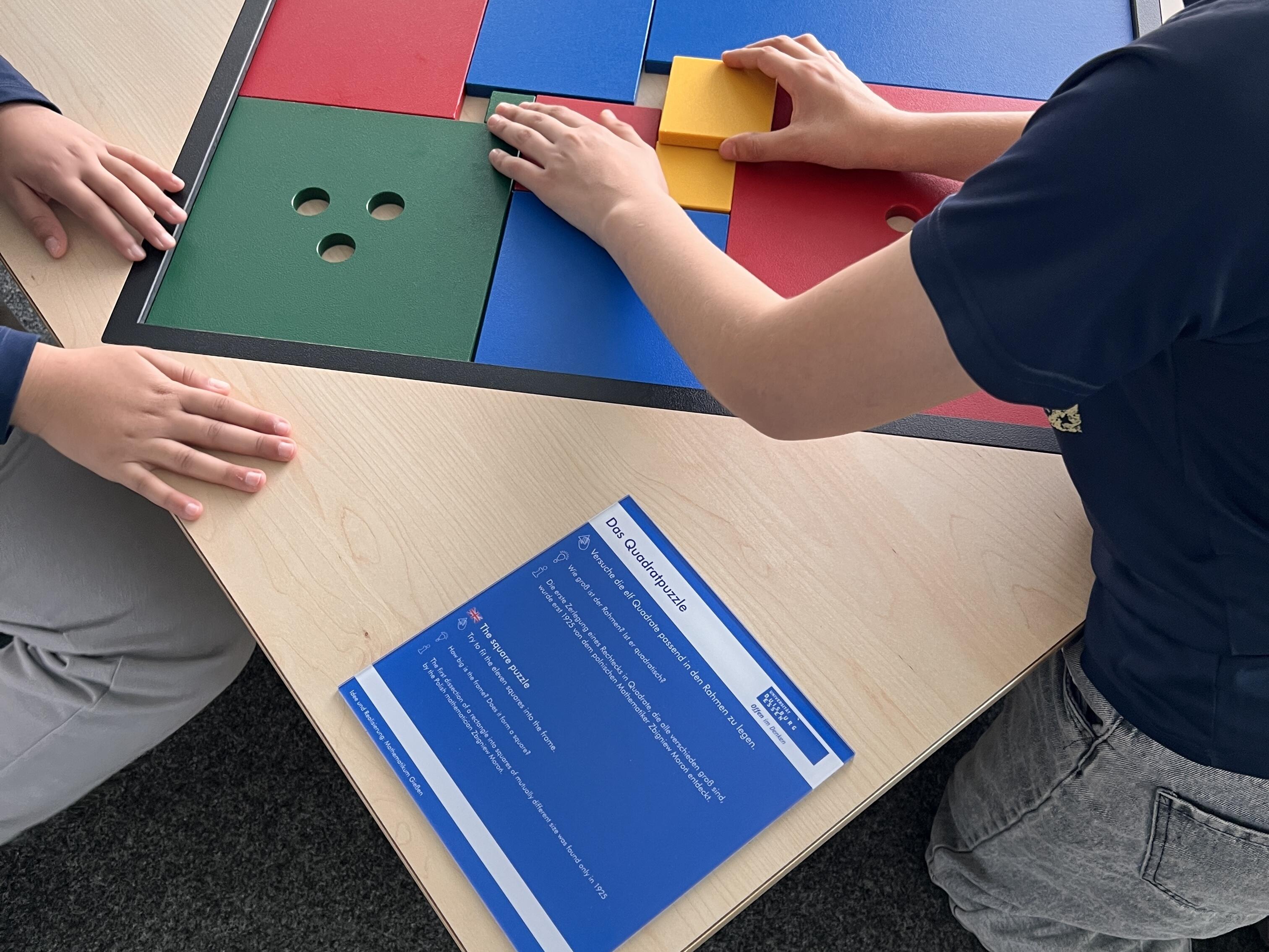 Kurzbeschreibung:
Zwei Kinder lösen gemeinsam ein großes Quadratpuzzle mit bunten Platten auf einem hellen Holztisch.

Ausführliche Bildbeschreibung:
Auf einem hellen Holztisch liegt ein großes quadratisches Spielfeld mit schwarzem Rahmen. Darin befinden sich verschieden große, farbige Platten in Gelb, Blau, Grün und Rot. Einige Platten haben mehrere runde Löcher.

Rechts sitzt ein Kind am Tisch und verschiebt mit beiden Händen ein gelbes Puzzleteil. Links sind die Hände eines weiteren Kindes zu sehen, das ebenfalls am Tisch steht oder sitzt. Vor dem Spielfeld liegt eine blaue Informationskarte mit der Überschrift „Das Quadratpuzzle“.

Das Bild zeigt eine interaktive Mitmachstation, bei der Kinder geometrische Formen anordnen und gemeinsam eine passende Lösung suchen. Der Fokus liegt auf dem spielerischen Ausprobieren, Sortieren und Kombinieren der farbigen Flächen.

(Bildbeschreibung mit KI generiert.)
