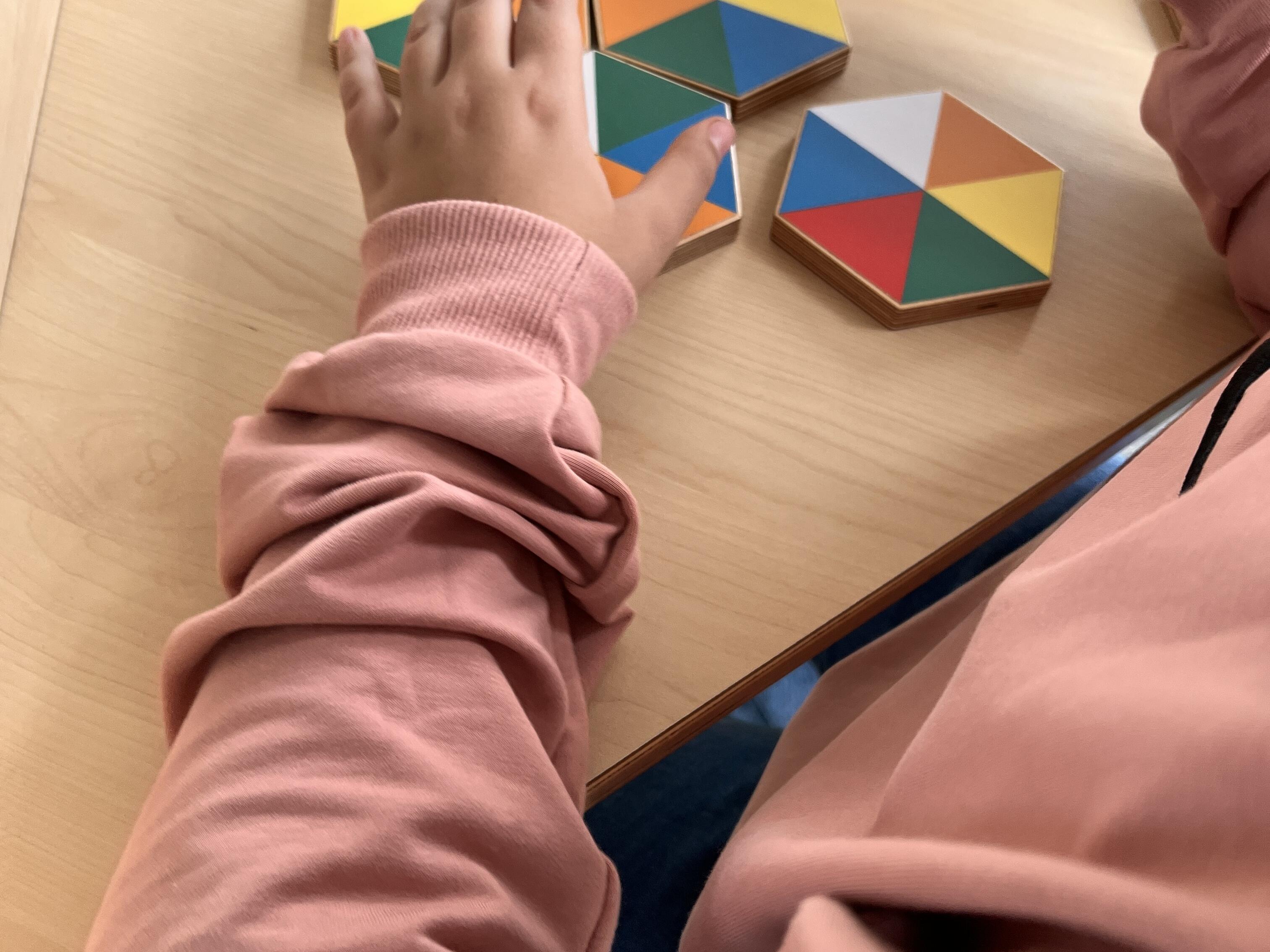 Kurzbeschreibung:
Ein Kind in einem rosa Pullover ordnet bunte sechseckige Holzplättchen auf einem hellen Holztisch an.

Ausführliche Bildbeschreibung:
Auf einem hellen Holztisch liegen mehrere sechseckige Holzplättchen. Jedes Plättchen ist in farbige Dreiecke unterteilt, unter anderem in Rot, Blau, Gelb, Grün, Orange und Weiß.

Ein Kind im rosa Pullover sitzt oder steht direkt am Tisch und bewegt die Plättchen mit beiden Händen. Einige Plättchen liegen einzeln, andere sind dicht beieinander angeordnet. Im oberen Bildbereich ist eine blaue Informationskarte teilweise zu sehen.

Das Bild zeigt eine interaktive Mitmachstation oder ein Lernspiel, bei dem Muster, Farben und Formen kombiniert werden. Der Fokus liegt auf dem spielerischen Ausprobieren und Ordnen der geometrischen Teile.

(Bildbeschreibung mit KI generiert.)