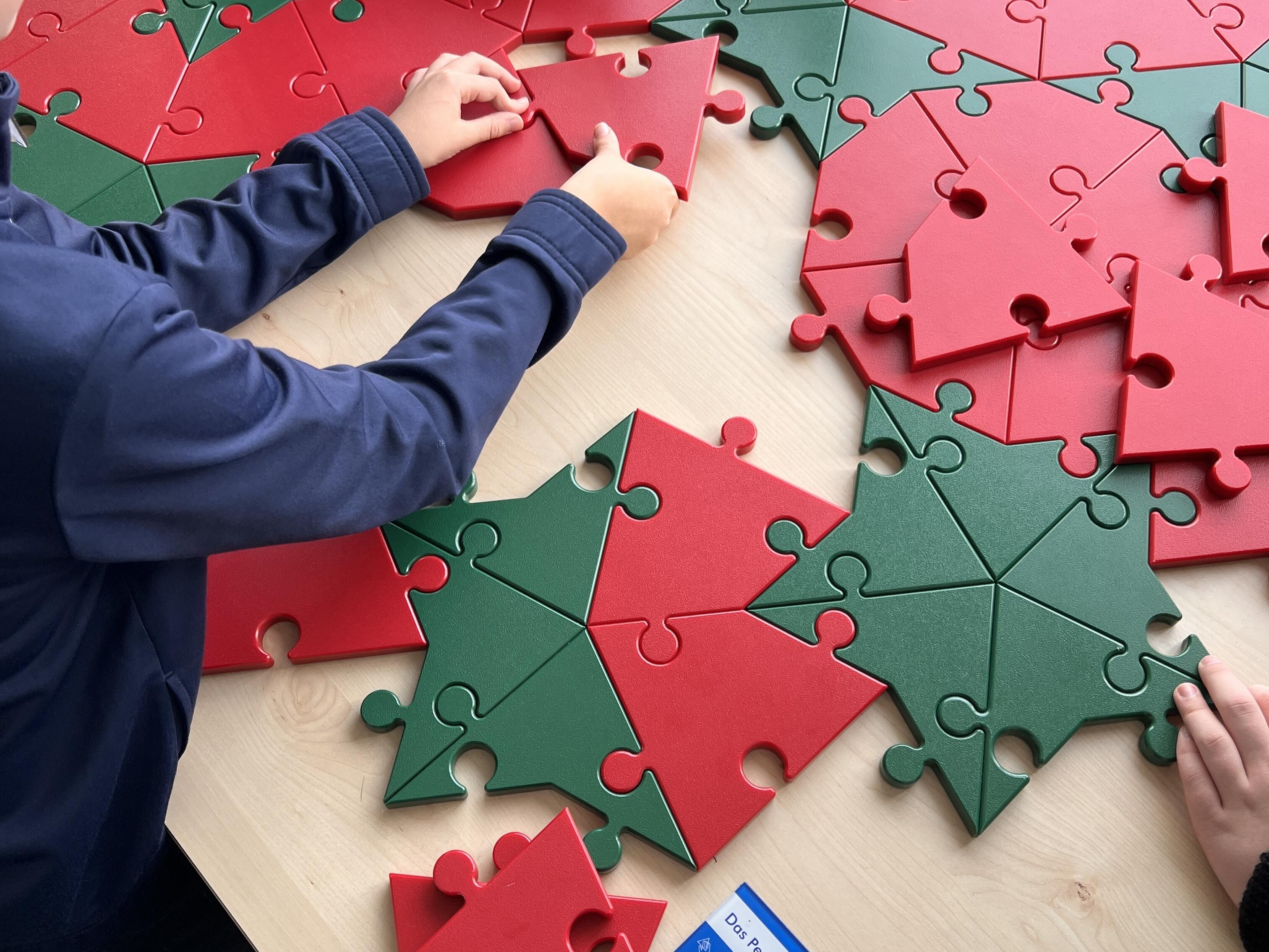 Kurzbeschreibung:
Mehrere Kinder setzen rote und grüne Puzzleteile auf einem hellen Holztisch zusammen. Die Teile bilden geometrische Muster aus stern- und hausähnlichen Formen.

Ausführliche Bildbeschreibung:
Auf einem hellen Holztisch liegen viele große Puzzleteile in Rot und Grün. Die Teile haben abgerundete Steckverbindungen und eingearbeitete Dreiecksformen. Einige Puzzleteile sind bereits zu größeren Mustern verbunden, andere liegen lose daneben.

Mehrere Kinder arbeiten gleichzeitig an dem Puzzle. Zu sehen sind vor allem Hände und Arme in blauen Pullovern, die einzelne Teile halten, verschieben oder an passende Stellen legen. Unten auf dem Tisch liegt eine blaue Informationskarte mit der Überschrift „Das Penrose-Puzzle“.

Das Bild zeigt eine interaktive Mitmachstation, bei der Kinder gemeinsam geometrische Formen zusammensetzen. Der Fokus liegt auf dem gemeinsamen Ausprobieren, Vergleichen und Finden passender Verbindungen.

(Bildbeschreibung mit KI generiert.)