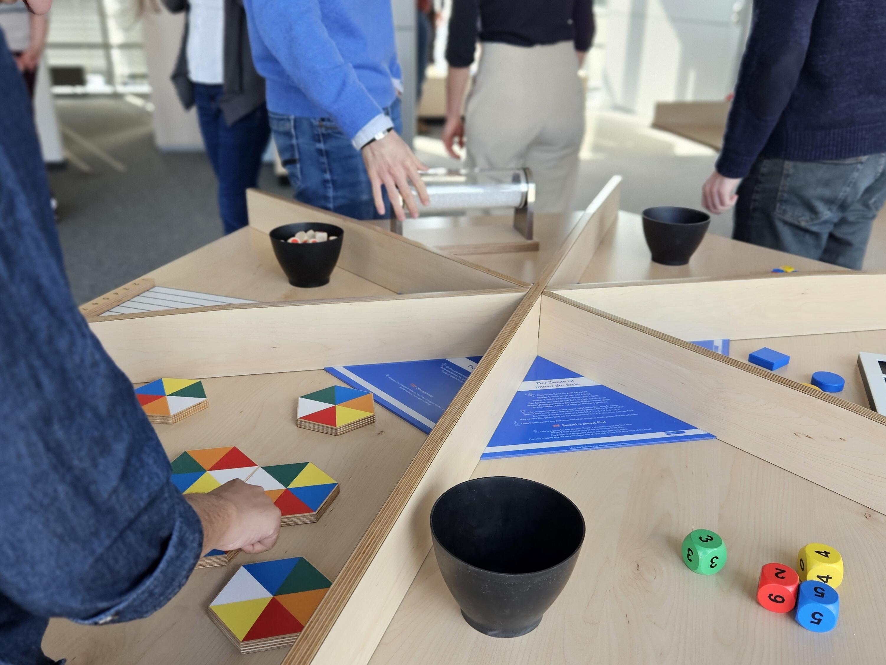 Kurzbeschreibung:
Mehrere Personen stehen an einem großen, sternförmig unterteilten Holztisch und beschäftigen sich mit bunten geometrischen Spiel- und Lernmaterialien. Auf den einzelnen Tischsegmenten liegen farbige Holzplättchen, Formen, Würfel und Schalen.

Ausführliche Bildbeschreibung:
Das Foto zeigt eine helle Innenraumsituation mit mehreren Personen, die sich um einen großen, sternförmig in sechs Segmente unterteilten Holztisch versammelt haben. Die Personen sind überwiegend von hinten oder seitlich zu sehen; Gesichter sind nicht erkennbar.

Im Vordergrund links legt eine Person ihre Hand auf ein sechseckiges Holzpuzzle mit bunten dreieckigen Segmenten in Blau, Grün, Gelb, Rot, Orange und Weiß. Auf dem Tisch befinden sich weitere gleichartige Holzplättchen mit unterschiedlichen Farbmustern.

Im rechten Vordergrund stehen eine schwarze Schale sowie mehrere große, farbige Spielwürfel mit aufgedruckten Zahlen. Weiter rechts liegen verschiedene geometrische Kunststoffformen in Rot, Blau, Gelb und Grün auf einem Sortierbrett mit quadratischen Vertiefungen.

In den übrigen Tischsegmenten befinden sich weitere schwarze Schalen sowie verschiedene Materialien. In der Tischmitte ist eine transparente zylindrische Vorrichtung auf einem Holzgestell platziert. Außerdem liegen blaue Informationsbroschüren auf dem Tisch.

Der Raum ist modern eingerichtet und wird durch große Fenster mit Tageslicht beleuchtet. Die Atmosphäre wirkt wie ein Workshop, eine Ausstellung oder eine interaktive Lernstation, an der mehrere Personen gemeinsam Materialien ausprobieren.

(Bildbeschreibung mit KI erstellt.)