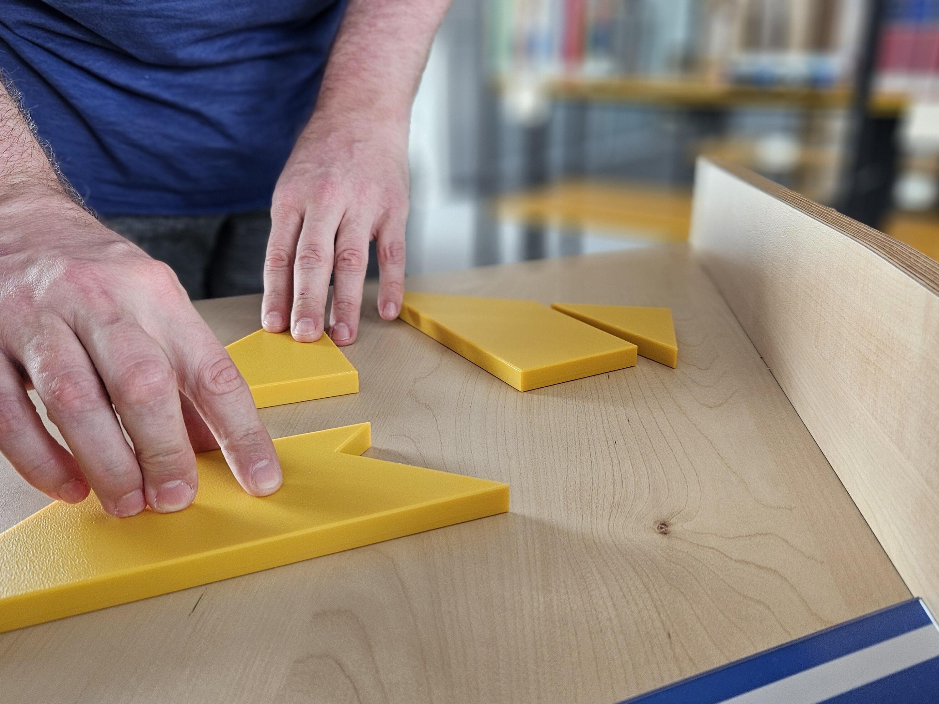 Gelbe Holztafeln in verschiedenen geoemtrischen Formen werden auf einem Holztisch von Händen verschoben. Es handelt sich dabei um Mitmach-Exponate. Inhaltlicher Hintegrund: Wer mathematische Themen buchstäblich anfassen und ausprobieren kann, versteht Zusammenhänge oft leichter. Die gelben Holztafeln zum Beispiel lassen sich leichter zum »T« zusammenfügen, wenn man sich auf die Eigenschaften der zu legenden Figur fokussiert und vergegenwärtigt, wie rechte Winkel gelegt werden können. Und wer das Rätsel schließlich erfolgreich gelöst hat, hat vielleicht auch ganz nebenbei Berührungsängste vor der Mathematik vergessen.