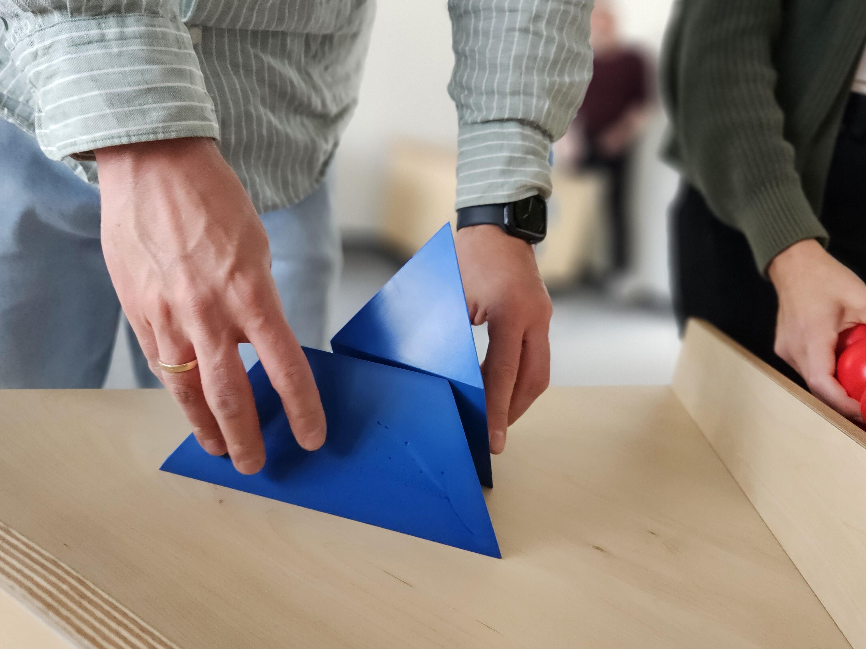 Kurzbeschreibung:
Zwei Personen stehen an einem hellen Holztisch und arbeiten mit einem blauen geometrischen Körper. Rechts liegt eine Holzkiste mit roten Kugeln.

Ausführliche Bildbeschreibung:
Im Vordergrund sind die Hände zweier Personen zu sehen. Eine Person setzt oder hält einen blauen geometrischen Körper aus mehreren dreieckigen Flächen auf einem hellen Holztisch. Die zweite Person steht rechts daneben und greift in eine Holzkiste mit mehreren roten Kugeln.

Die Gesichter der Personen sind nicht zu sehen, der Fokus liegt auf den Händen und den Objekten. Der Hintergrund ist unscharf und lässt einen hellen Innenraum erkennen. Das Bild zeigt eine Mitmachstation, an der spielerisch Formen und Materialien gemeinsam ausprobiert werden.

(Bildbeschibung mi KI generiert.)