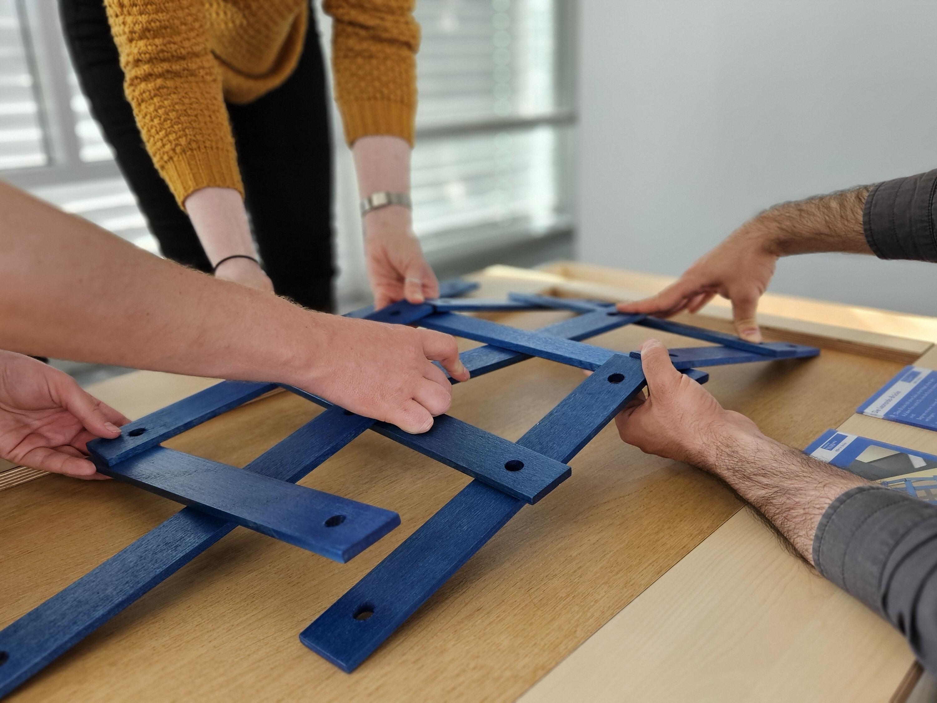 Kurzbeschreibung:
Mehrere Personen bauen gemeinsam an einem blauen Holzgestell auf einem hellen Tisch. Zu sehen sind vor allem Hände, die blaue Leisten halten und anordnen.

Ausführliche Bildbeschreibung:
Auf einem hellen Holztisch liegen mehrere blaue Holzleisten mit runden Löchern. Mehrere Personen stehen um den Tisch herum und halten die Leisten mit ihren Händen fest. Sie scheinen gemeinsam ein geometrisches Muster oder eine Konstruktion zusammenzusetzen.

Die Personen sind nur teilweise sichtbar: Man sieht Arme, Hände und Oberkörper, aber keine Gesichter. Eine Person trägt einen gelben Pullover, andere tragen dunkle oder graue Kleidung. Rechts auf dem Tisch liegen blaue Informationskarten oder Anleitungen.

Der Hintergrund ist leicht unscharf und zeigt einen hellen Innenraum mit großen Fenstern. Das Bild vermittelt den Eindruck einer interaktiven Mitmachstation, bei der Menschen gemeinsam ausprobieren, konstruieren und zusammenarbeiten.

(Bildbeschreibung mit KI generiert.)
