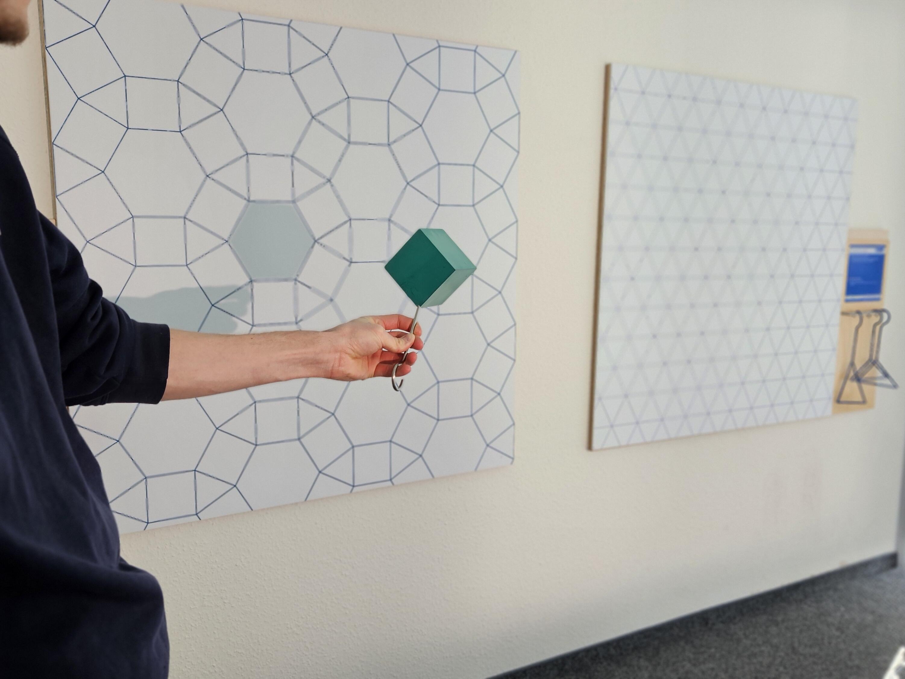Kurzbeschreibung:
Eine Person hält ein grünes, rautenförmiges Element vor eine Wandtafel mit einem geometrischen Linienmuster. Im Hintergrund sind eine weitere Musterfläche und ein heller Innenraum mit Fenstern zu sehen.

Ausführliche Bildbeschreibung:
Das Foto zeigt einen hellen Innenraum mit zwei großen, an der Wand befestigten Tafeln. Beide Tafeln sind mit regelmäßigen geometrischen Linienmustern versehen. Die linke Tafel zeigt ein Netz aus Sechs- und Vierecken, während die rechte Tafel ein Dreiecksraster aufweist.

Links im Bild ist eine Person teilweise zu sehen. Ihr Oberkörper und ein ausgestreckter Arm sind erkennbar, das Gesicht ist nur angeschnitten. Die Person hält mit einer Zange oder einem Greifwerkzeug ein grünes, rautenförmiges dreidimensionales Element vor die linke Tafel. In der Mitte dieser Tafel befindet sich ein hellgrün markiertes Sechseck, das sich farblich vom übrigen Muster abhebt.

Rechts daneben hängt die zweite Tafel mit einem feinen Dreiecksmuster. Davor befindet sich eine kleine Halterung mit Informationsmaterial und einem daran befestigten Drahtgestell.

Der Raum wird durch große Fenster auf der rechten Seite mit Tageslicht beleuchtet. Der Boden ist mit grauem Teppich ausgelegt. Insgesamt vermittelt die Szene den Eindruck einer interaktiven Lern- oder Ausstellungsstation, an der geometrische Formen und Muster erkundet werden können.

(Bildbeschreibung mit KI erstellt.)