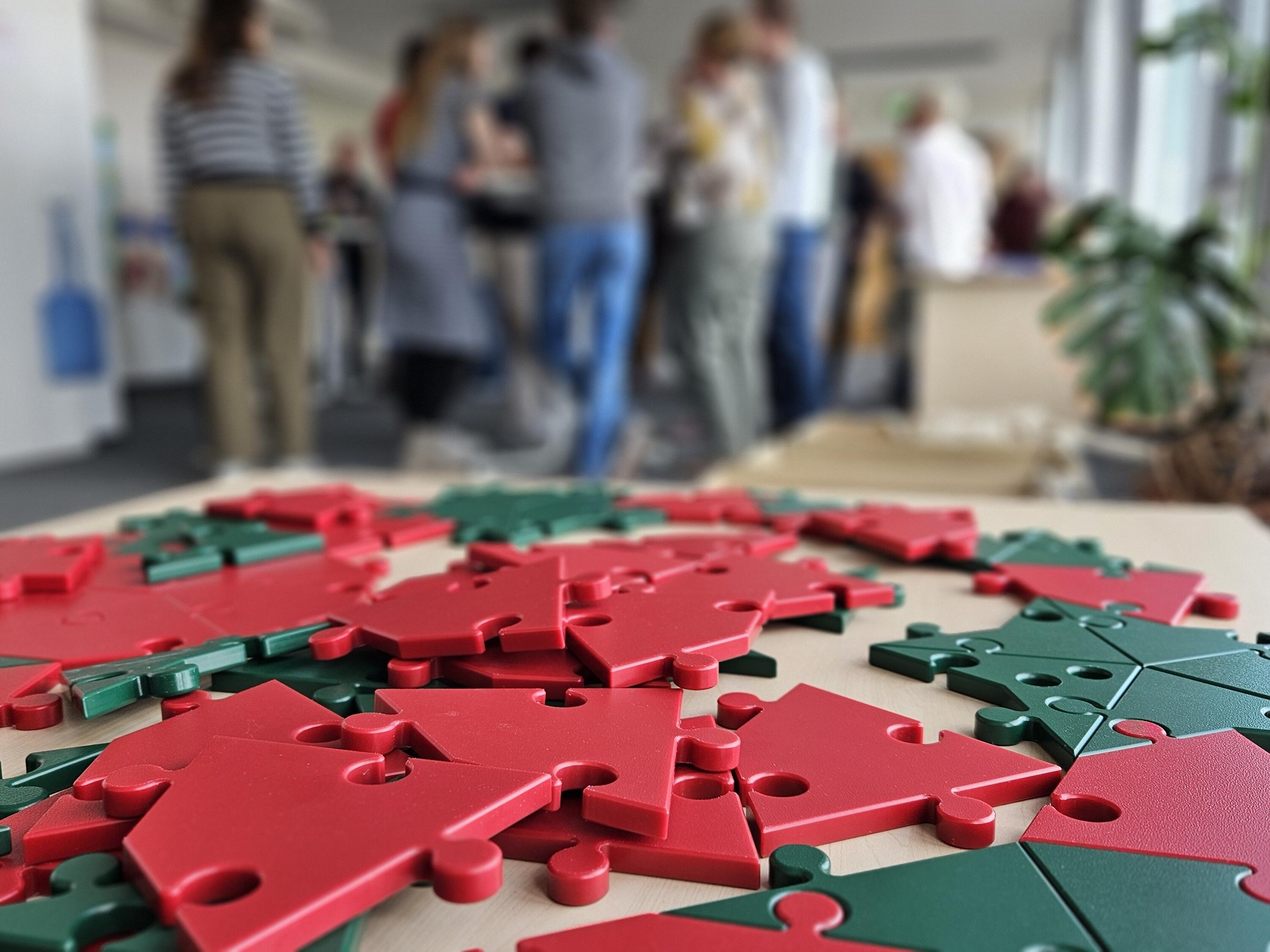 Kurzbeschreibung:
Auf einem Tisch liegen rote und grüne, hausförmige Puzzleteile. Im unscharfen Hintergrund stehen mehrere Menschen in einem hellen Raum beisammen.

Ausführliche Bildbeschreibung:
Im Vordergrund sind zahlreiche rote und grüne Kunststoff-Puzzleteile auf einem hellen Tisch ausgebreitet. Die Teile haben die Form kleiner Häuser und sind teilweise übereinandergelegt. Einige liegen bereits passend nebeneinander, andere sind lose verteilt.

Der Hintergrund ist bewusst unscharf: Dort stehen mehrere Personen in einem hellen Innenraum. Rechts sind große Fenster und eine grüne Zimmerpflanze zu erkennen. Durch die starke Tiefenschärfe liegt der Fokus deutlich auf den Puzzleteilen im Vordergrund. Das Bild vermittelt den Eindruck von gemeinschaftlichem Arbeiten, Austausch und Zusammenfügen einzelner Elemente zu einem größeren Ganzen.

(Bildbeschreibung mit KI erstellt.)