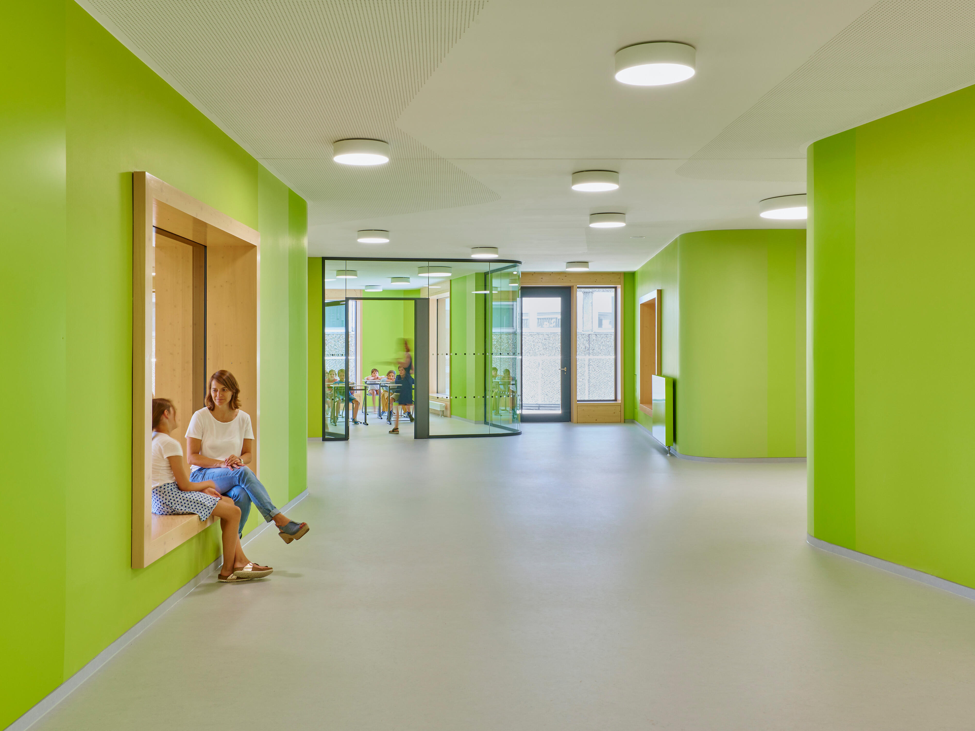 Heller, großzügiger Flur mit grün gefärbten Wänden rechts und links, kreisförmigen weißen Lampen an der hellen Decke. Links im Bild ist eine Holzvertäfelte Sitznische in die Wand eingelassen, in der eine weiblich gelesene erwachsene Person und ein weiblich gelesenes Kind sitzen und sich unterhalten.