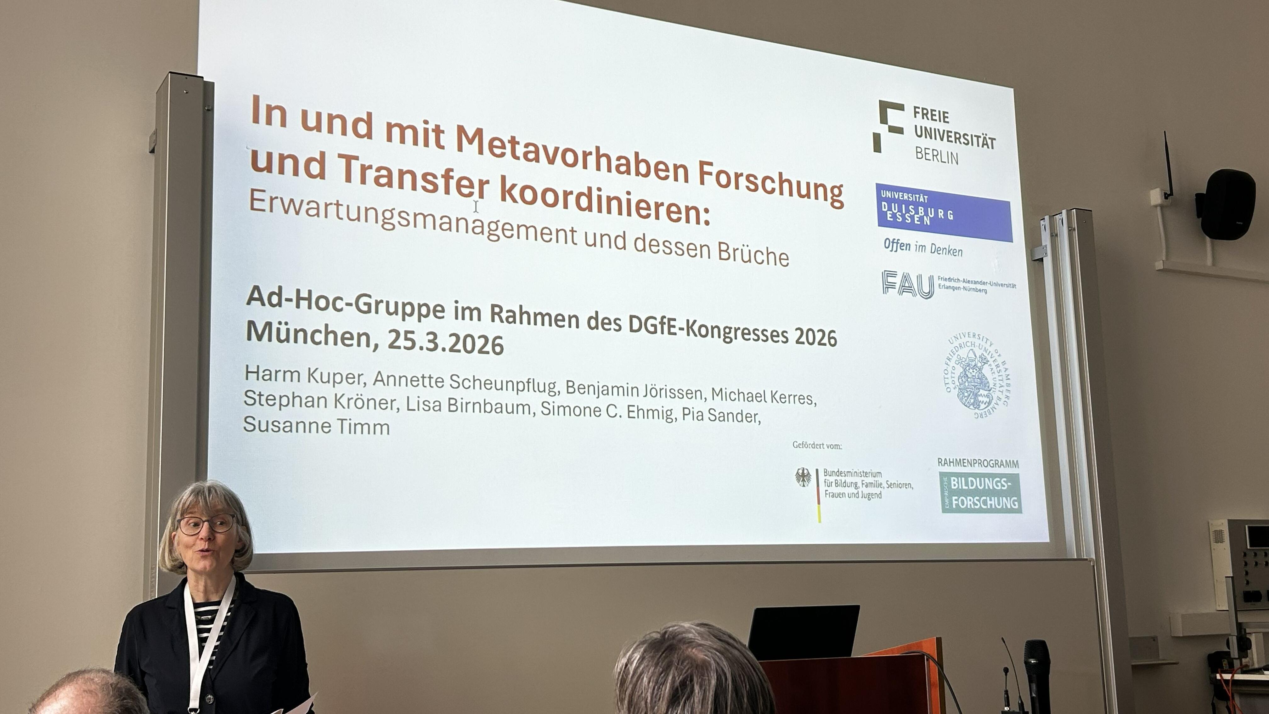 Eine Frau steht in einem Raum, im Hintergrund ist eine Präsentation an die Wand projiziert.