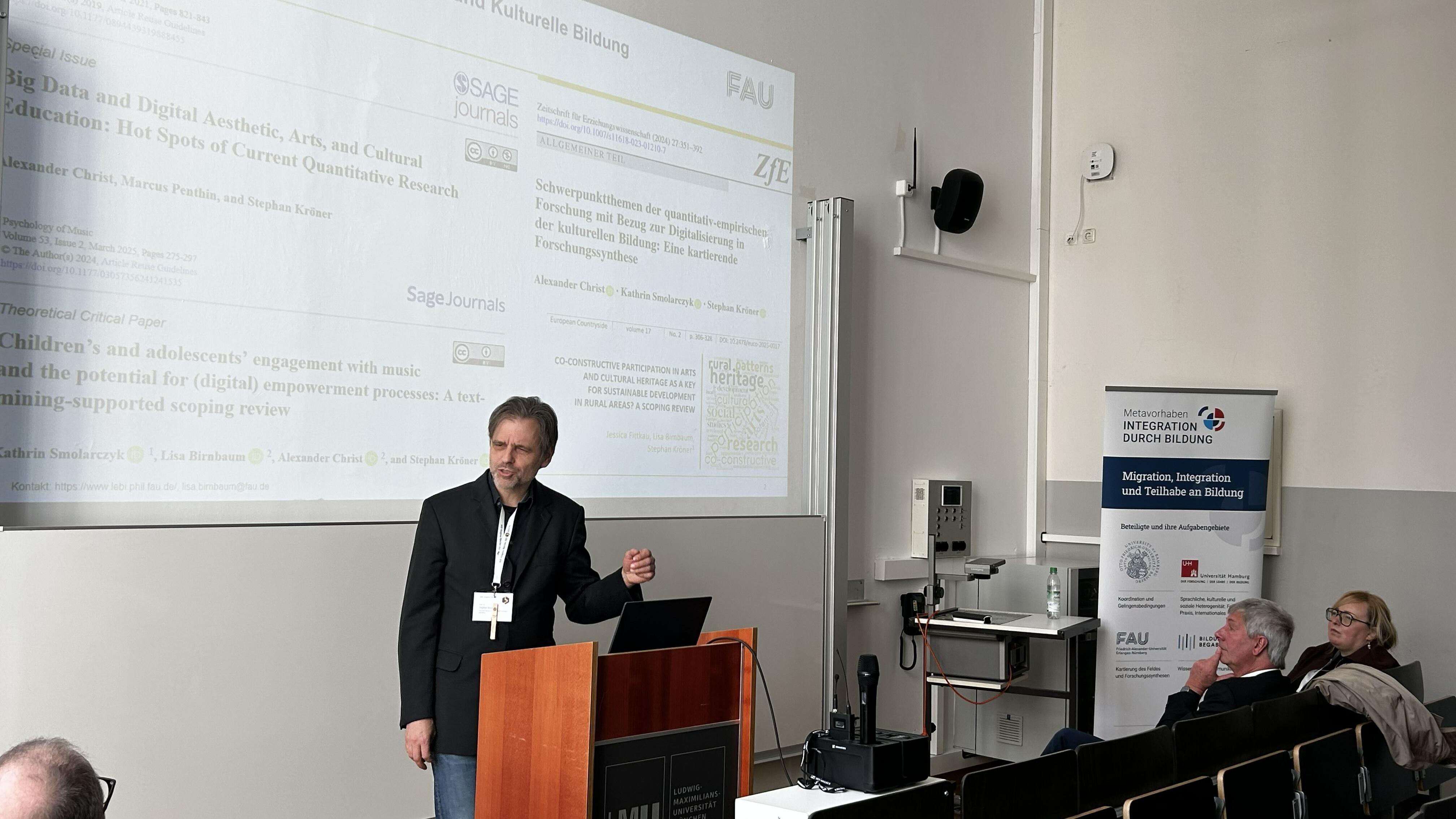 Ein Mann steht an einem Pult, im Hintergrund ist eine Präsentation an die Wand projiziert.