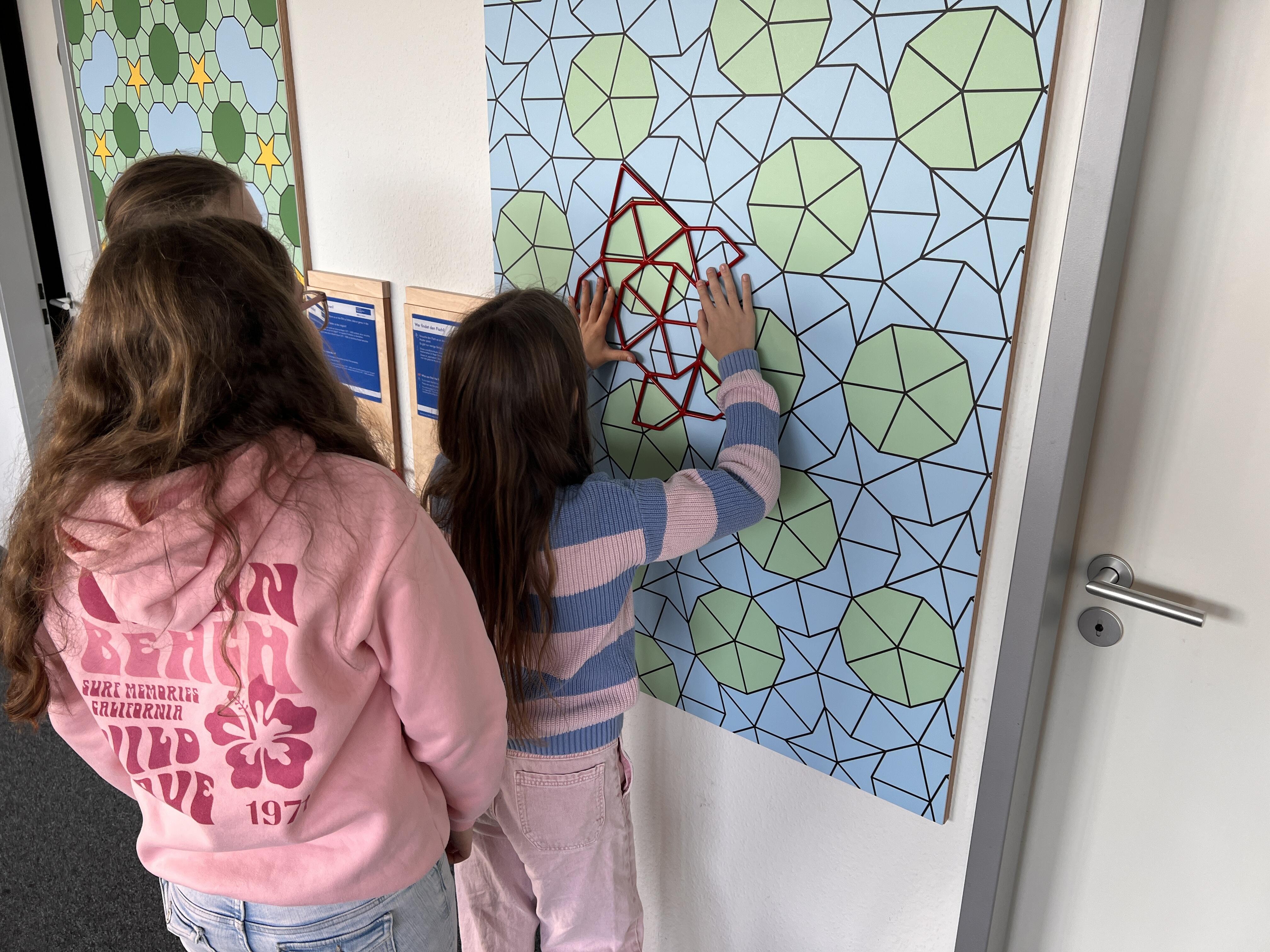 Kurzbeschreibung:
Drei Kinder stehen vor einer Wandtafel mit geometrischem Muster. Ein Kind legt eine rote Form auf das Muster an.

Ausführliche Bildbeschreibung:
Auf dem Bild sind drei Kinder von hinten beziehungsweise seitlich zu sehen. Sie stehen vor einer großen Wandtafel mit einem geometrischen Muster aus vielen schwarzen Linien, hellblauen Flächen und grünen Vielecken.

Ein Kind in einem gestreiften Pullover hält mit beiden Händen ein rotes, gitterartiges Formteil an die Tafel. Es scheint zu prüfen, wie die rote Form in das Muster passt. Zwei weitere Kinder stehen daneben und schauen zu. Links an der Wand sind weitere Tafeln mit ähnlichen Mustern und blaue Informationskarten zu sehen. Rechts befindet sich eine helle Tür.

Das Bild zeigt eine interaktive Mitmachstation, bei der Kinder geometrische Formen vergleichen, anlegen und Muster entdecken. Der Fokus liegt auf gemeinsamem Ausprobieren und räumlichem Denken.

(Bildbeschreibung mit KI generiert.)