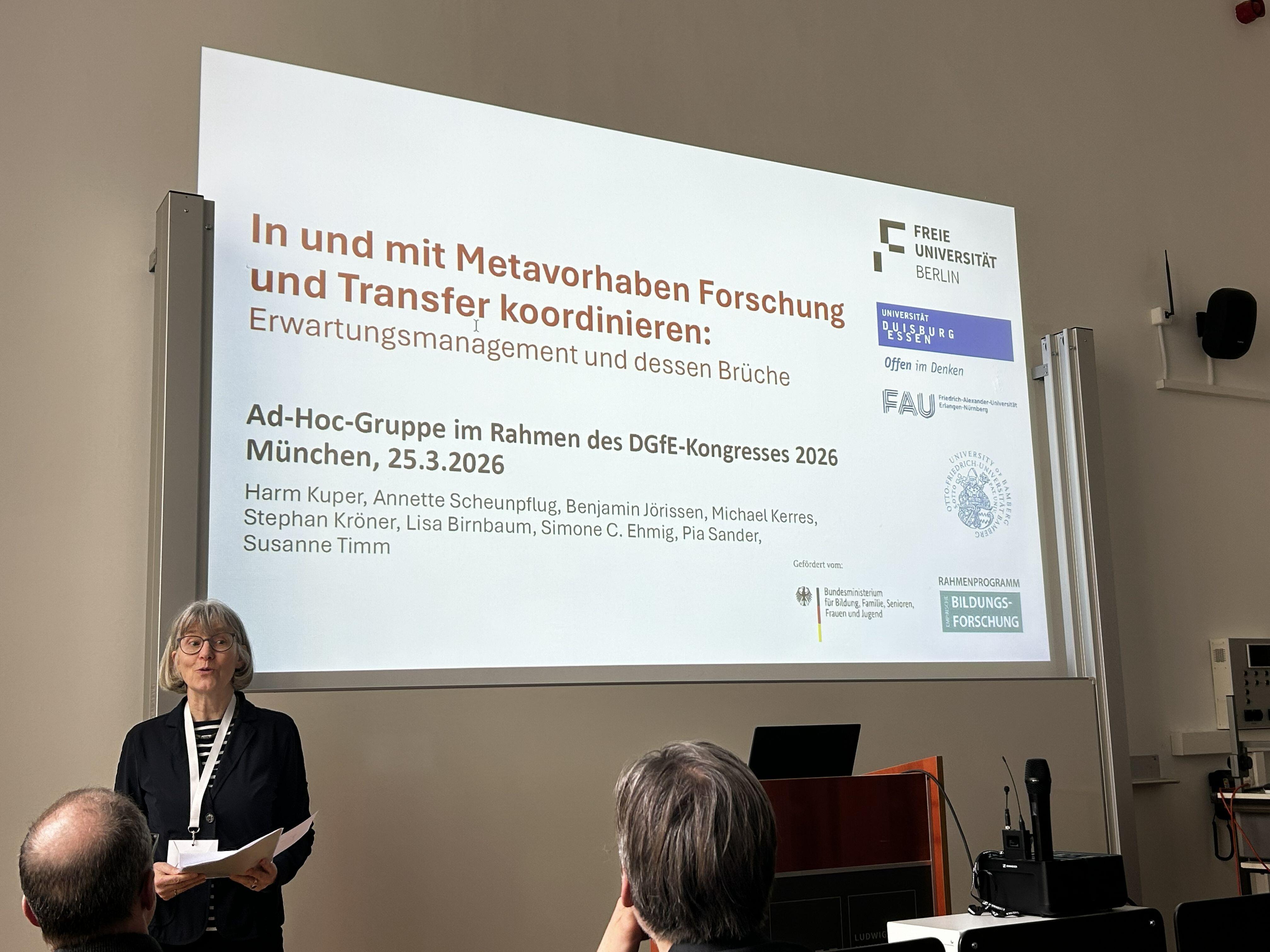 Eine Frau steht in einem Raum, im Hintergrund ist eine Präsentation an die Wand projiziert.