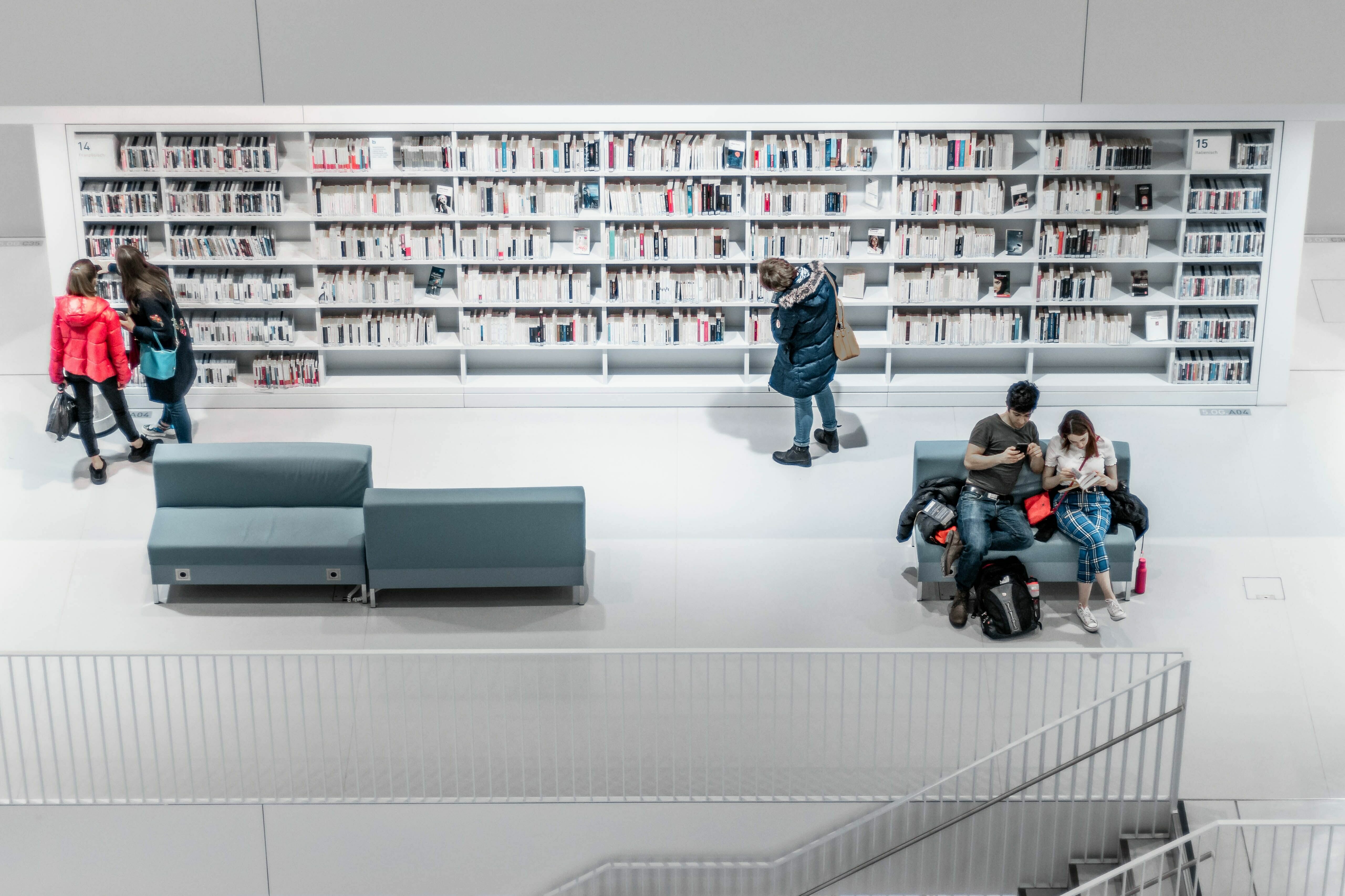 Abbildung einer Bibliothek mit Bücherregalen und Sitzecken sowie Personen, die lesen.