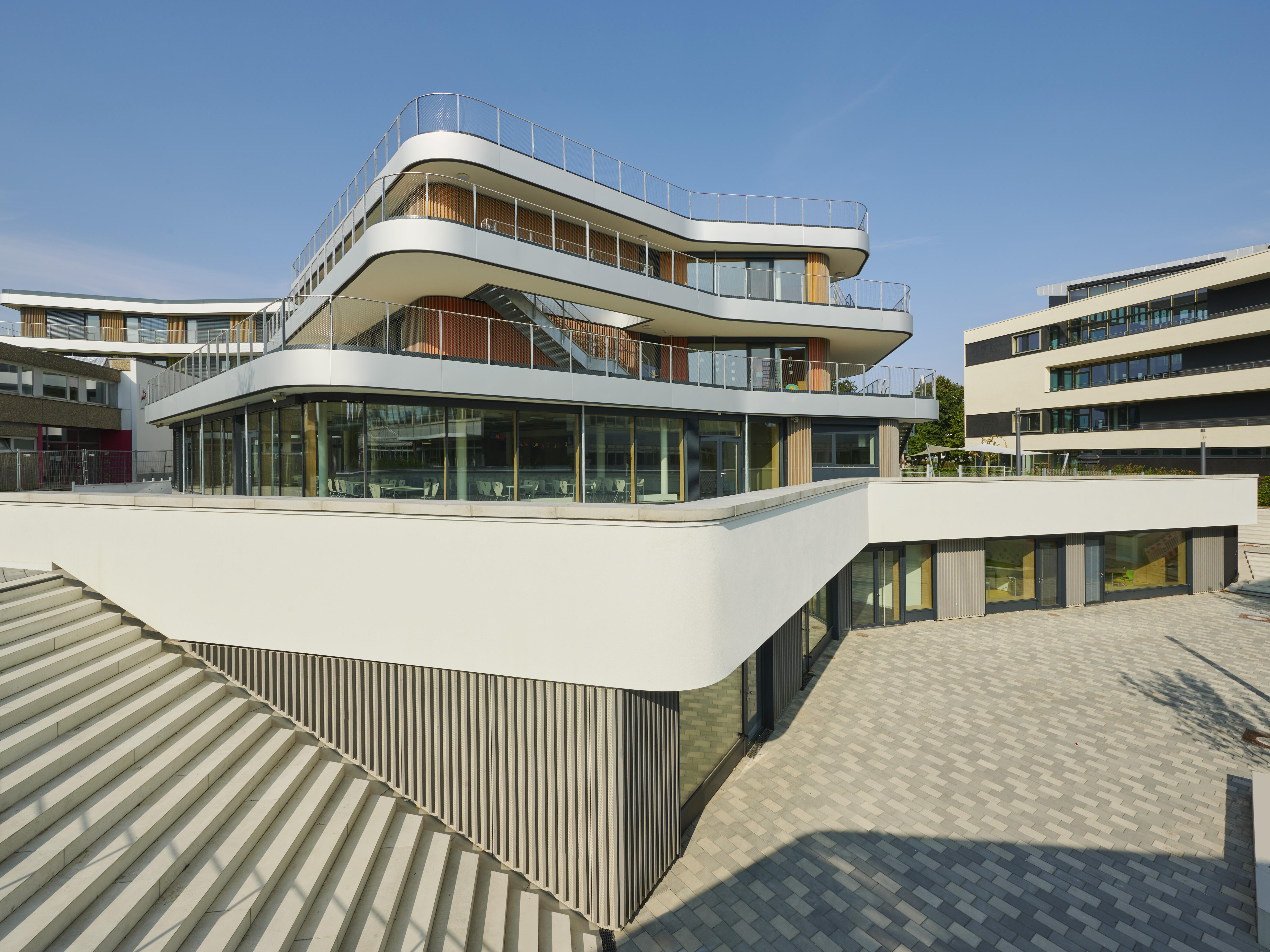 Schulhof mit Außenansicht der Gotthard-Müller-Grund- und Gemeinschaftsschule Filderstadt aus erhöhter Perspektive mit Panoramatreppe im linken Vordergrund. Im Hintegrund ist das Schulgebäude zu sehen, ein breiter, 4-geschossiger Flachdachbau, dessen Fassadenplatten gruppenweise in den Farben Gelb, Grün und Rot gefärbt sind.