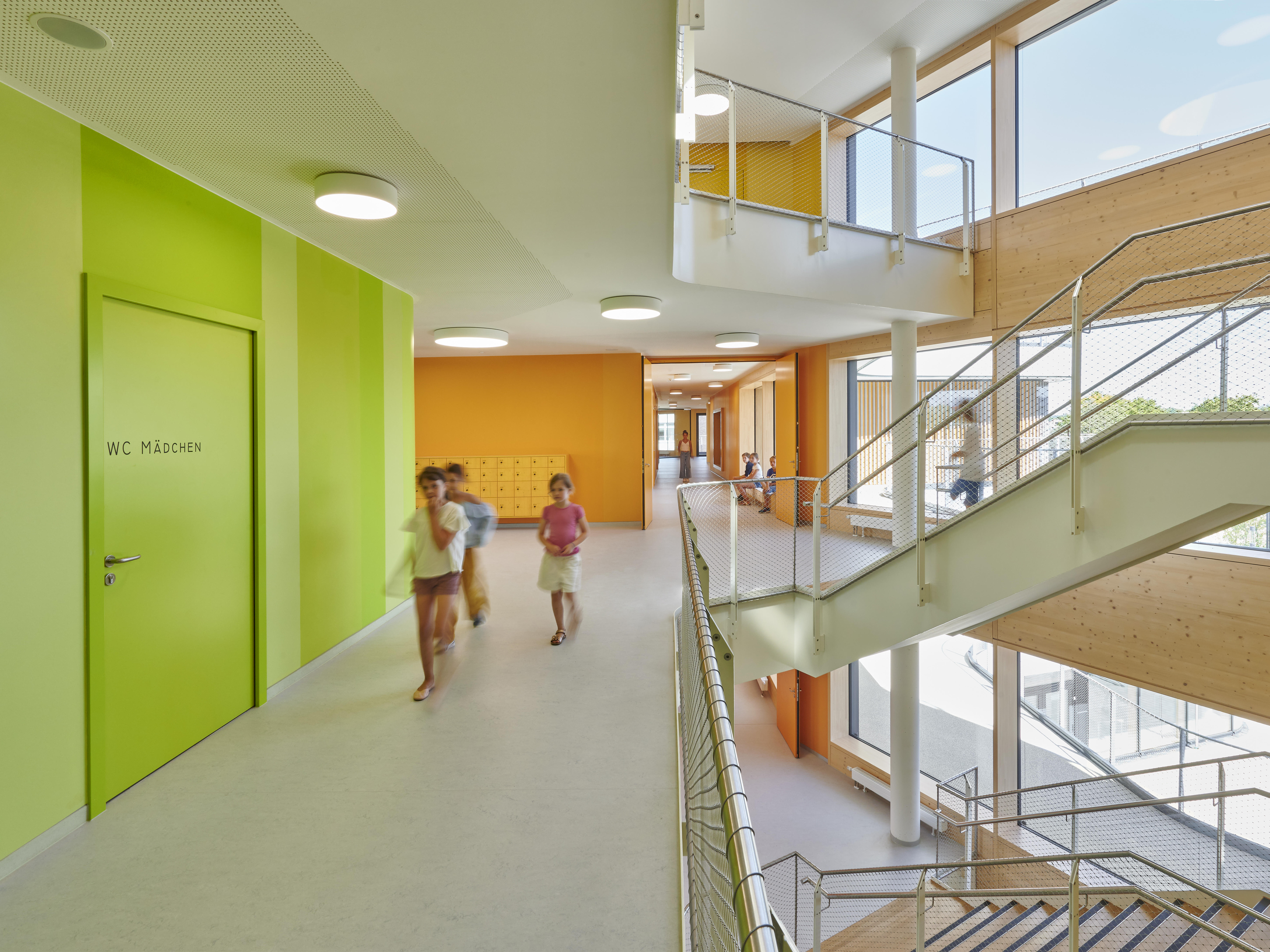 Halb offenes Treppenhaus der Gotthard-Müller-Schule ist eine Gemeinschaftsschule. Blick vom Gang der dritten Etage auf Treppen. Die darüber und darunter liegende Etage sind im Anschnitt zu sehen. Am linken Bildrand ist dominierend eine hell-grasgrüne Wand mit Tür in gleicher Farbe zu sehen. Im Hintergrund stößt der Blick auf durchgehend orangegefärbte Wände. Über den Gang kommen drei Kinder entgegen. Ihr Bild ist leicht verschwischt.