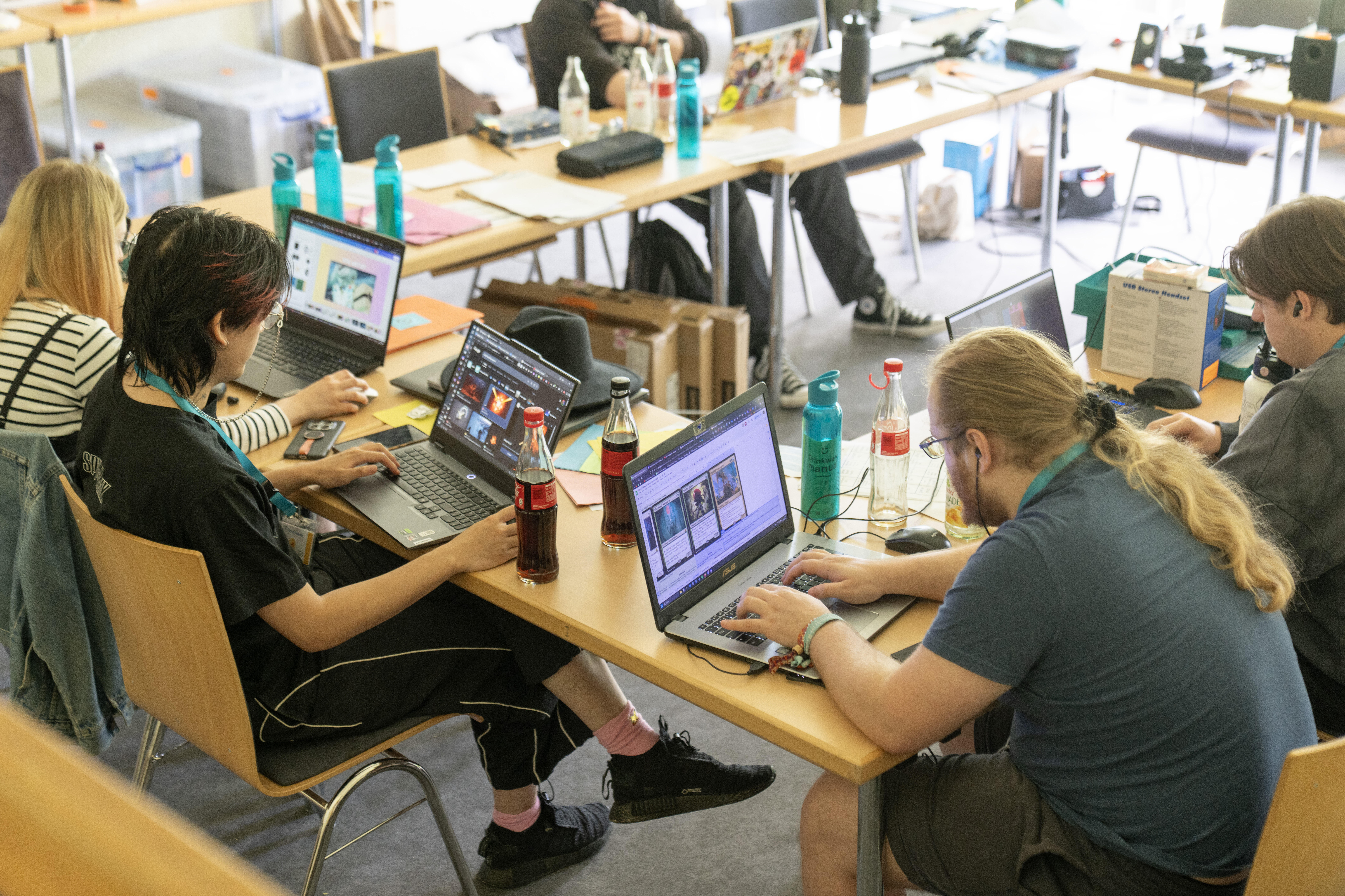 Jugendliche und Erwachsene sitzen locker an Laptops. Die Laptopbilschirme zeigen Gaming-Motive.