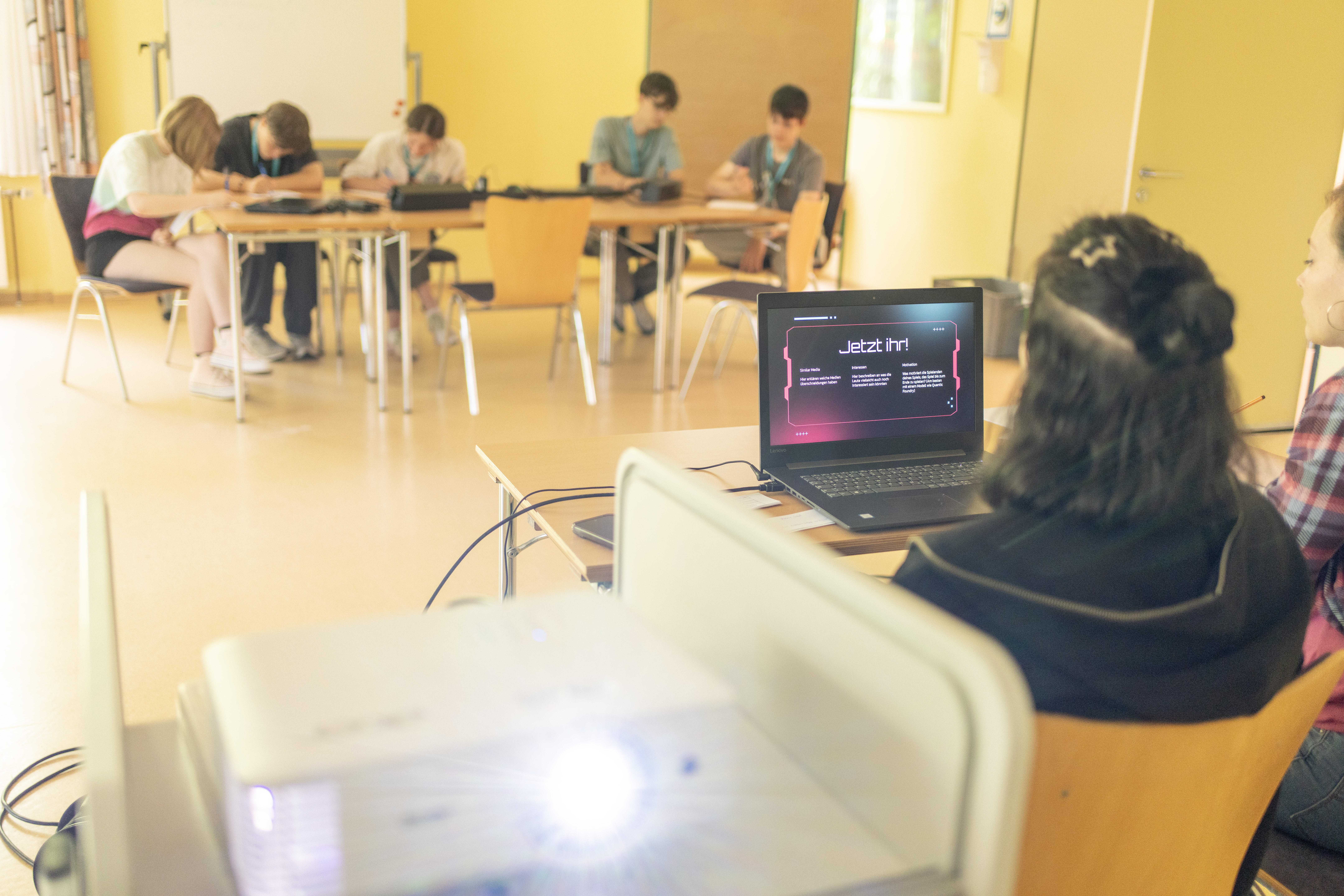 Schüler im Klassenraum in Arbeit an Laptops vertieft. Im Vordergrund Laptop-Monitor auf dem Präsentation läuft. Die Angezeigte Folie enthält die Aufforderung "Jetzt Ihr!"