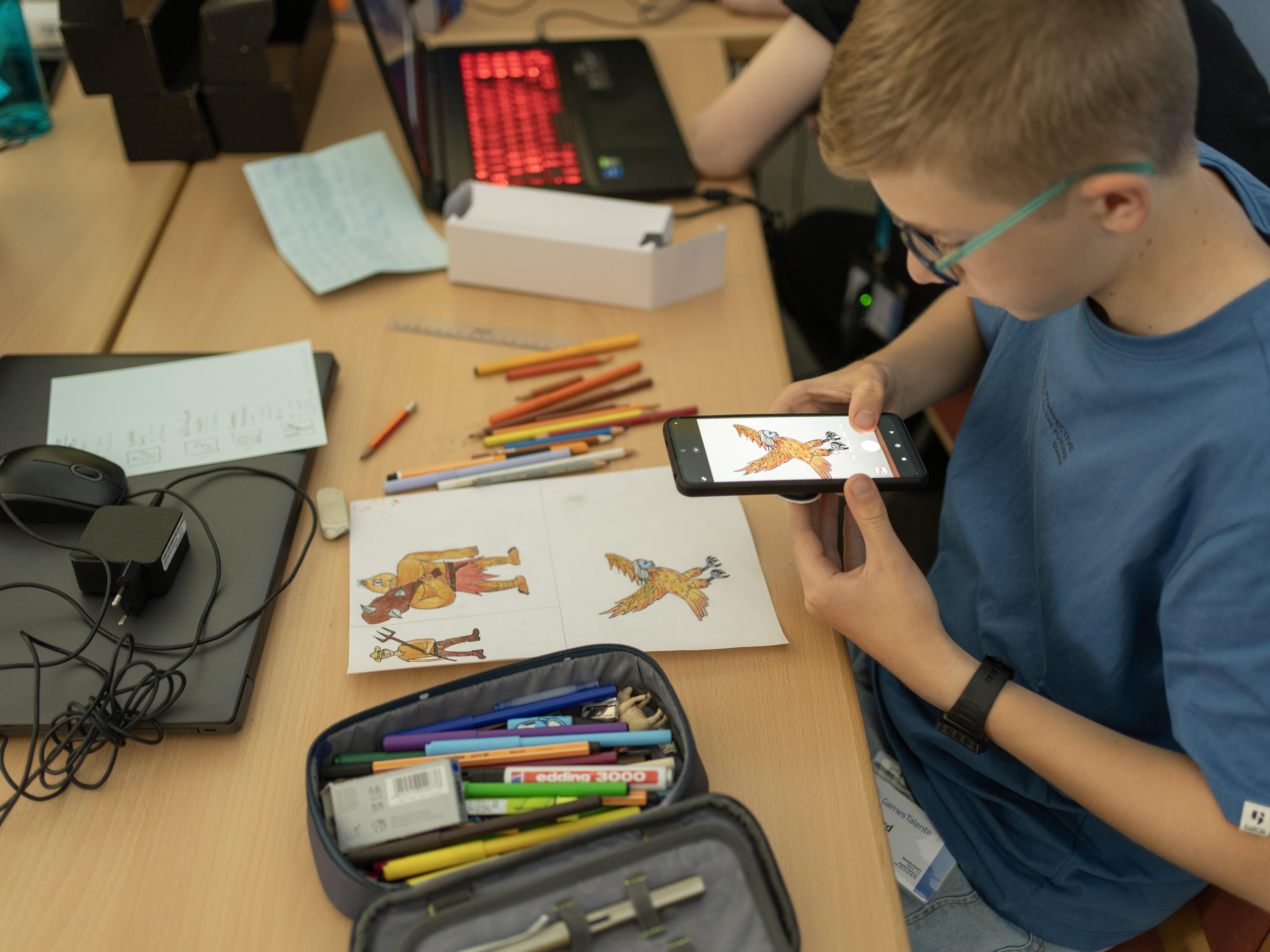 Ein Jugendlicher sitzt am Schreibtisch in einem Lehrraum und scannt mit einem Smartphone die colorierte Zeichnung eines Raubvogels. Auf dem Schreibtisch befindet sich ein geöffnetes Federmäppchen mit Buntstiften, die teils auch auf dem Tisch verteilt sind, sowie ein geschlossener Laptop mit Maus und Netztteil.