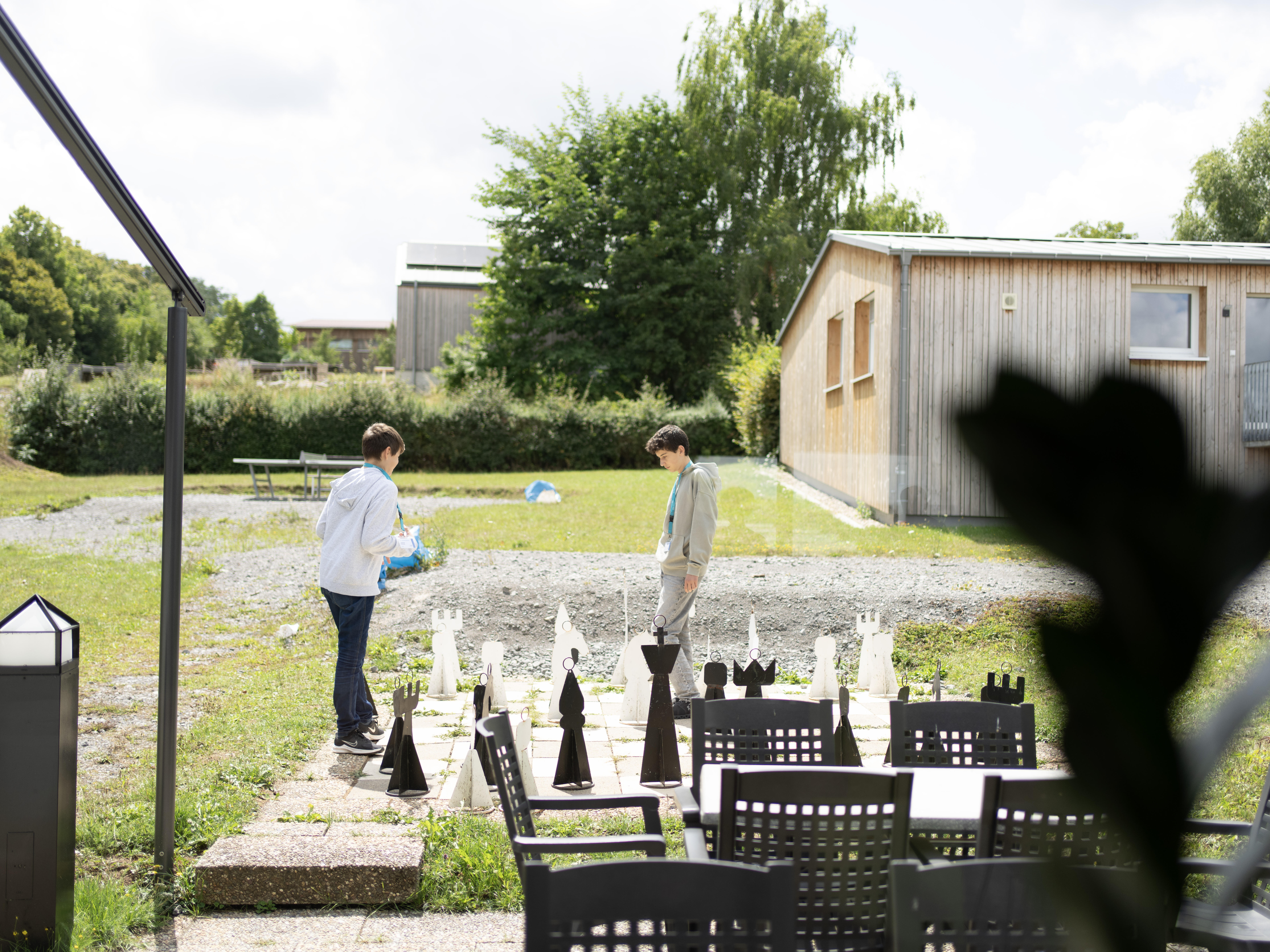Zwei Jugendliche spielen mit einem XXL-Outddoor-Schachspiel auf einer begrünten Schotter-Freifläche zwischen modernen Blockhütten. Im Vordergrund sieht man einen Gartentisch mit Stühlen.