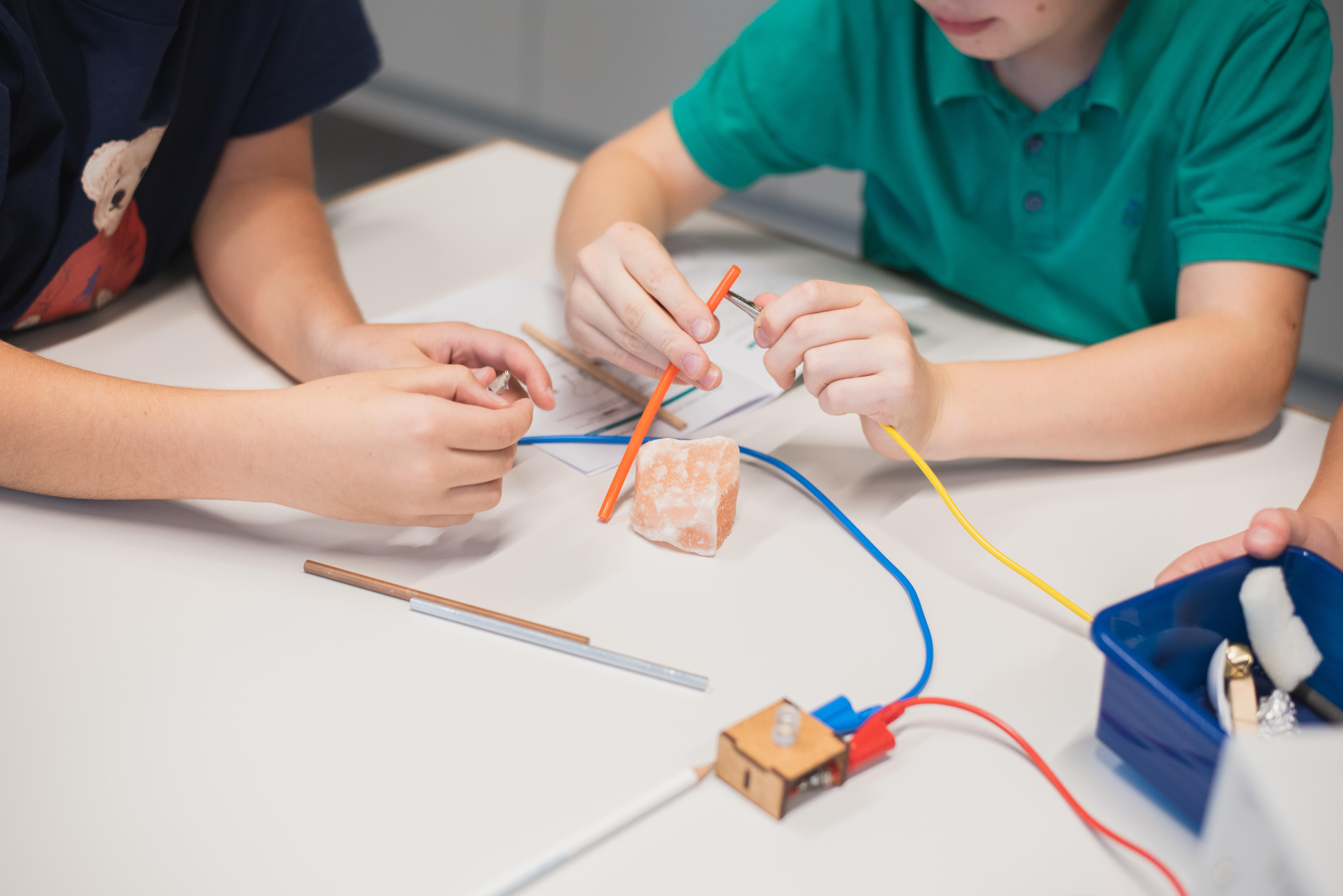 Ausschnitt eines Tisches mit Krokodilklemmenkabeln, einer LED und verschiedenen Materialien, Strohhalm, Stein, Holzstab. Zwei Kinder sitzen am Tisch. Eines schließt die Krokodilklemme an den Strohhalm an.