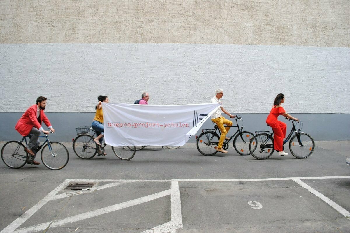 Abbildung von vier Fahrradfahrern, die seitlich ein Banner mit der Aufschrift "UNESCO-Projekts-Schulen" halten.