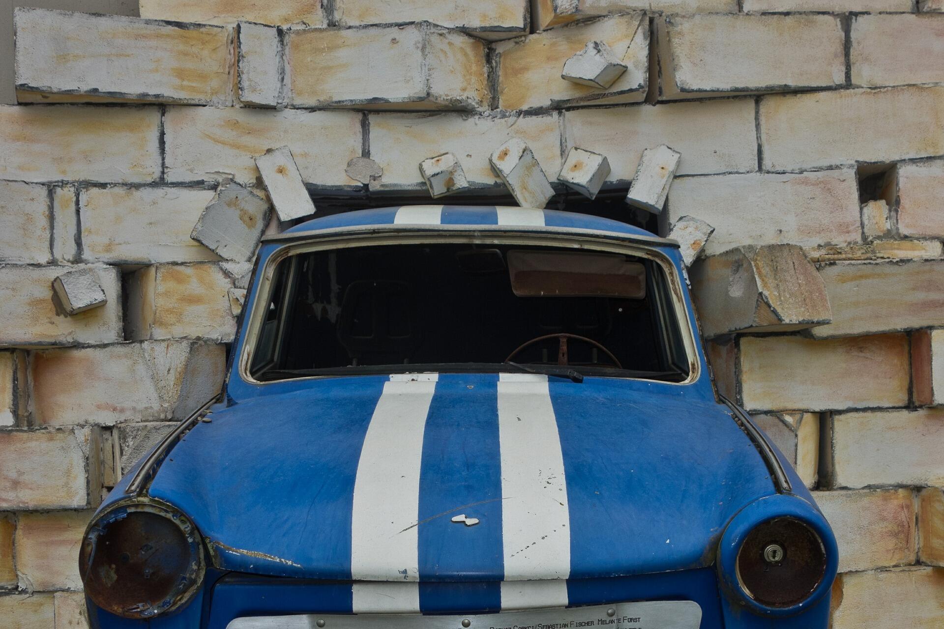 Blau-weiß gestreifter Trabi, der aus einer Mauer herausragt.