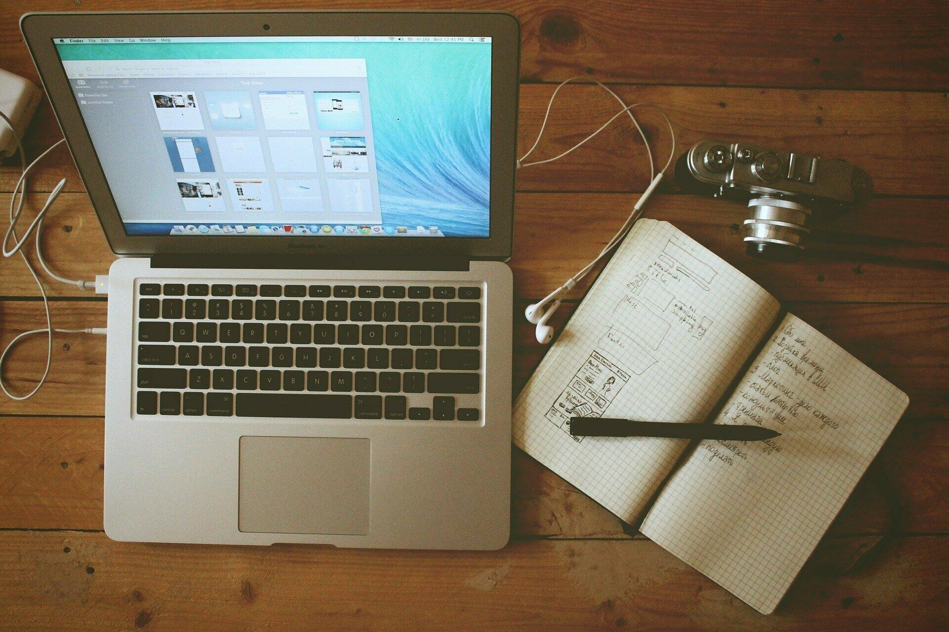 Laptop, Kamera, Heft und Stift auf einem Holztisch.