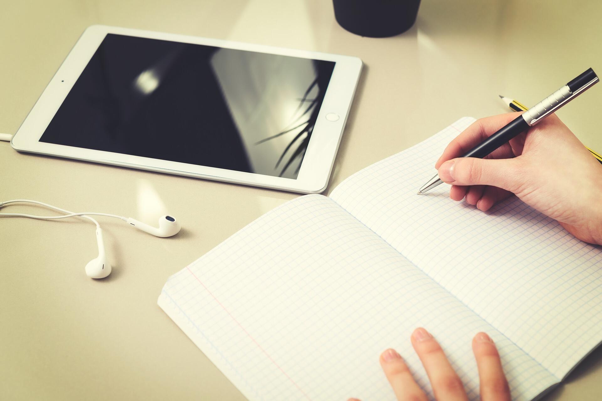 Tablet, Kopfhörer und Heft auf einem Tisch, dazu eine Person, die mit dem Stift etwas ins Heft schreiben will.