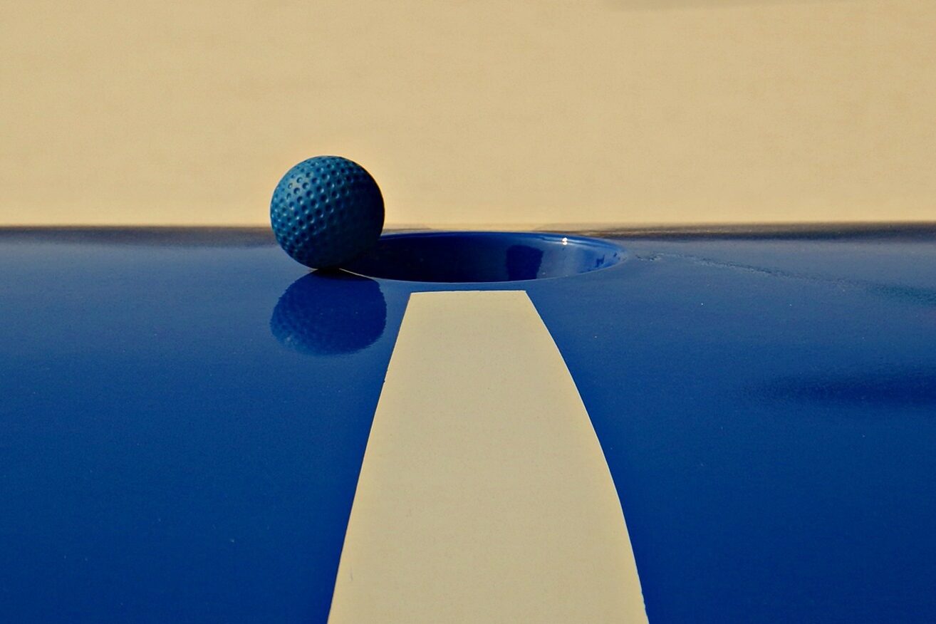 Abbildung einer blauen Minigolfbahn mit beigem Streifen und einem blauen Minigolf-Ball, der am Loch liegt.